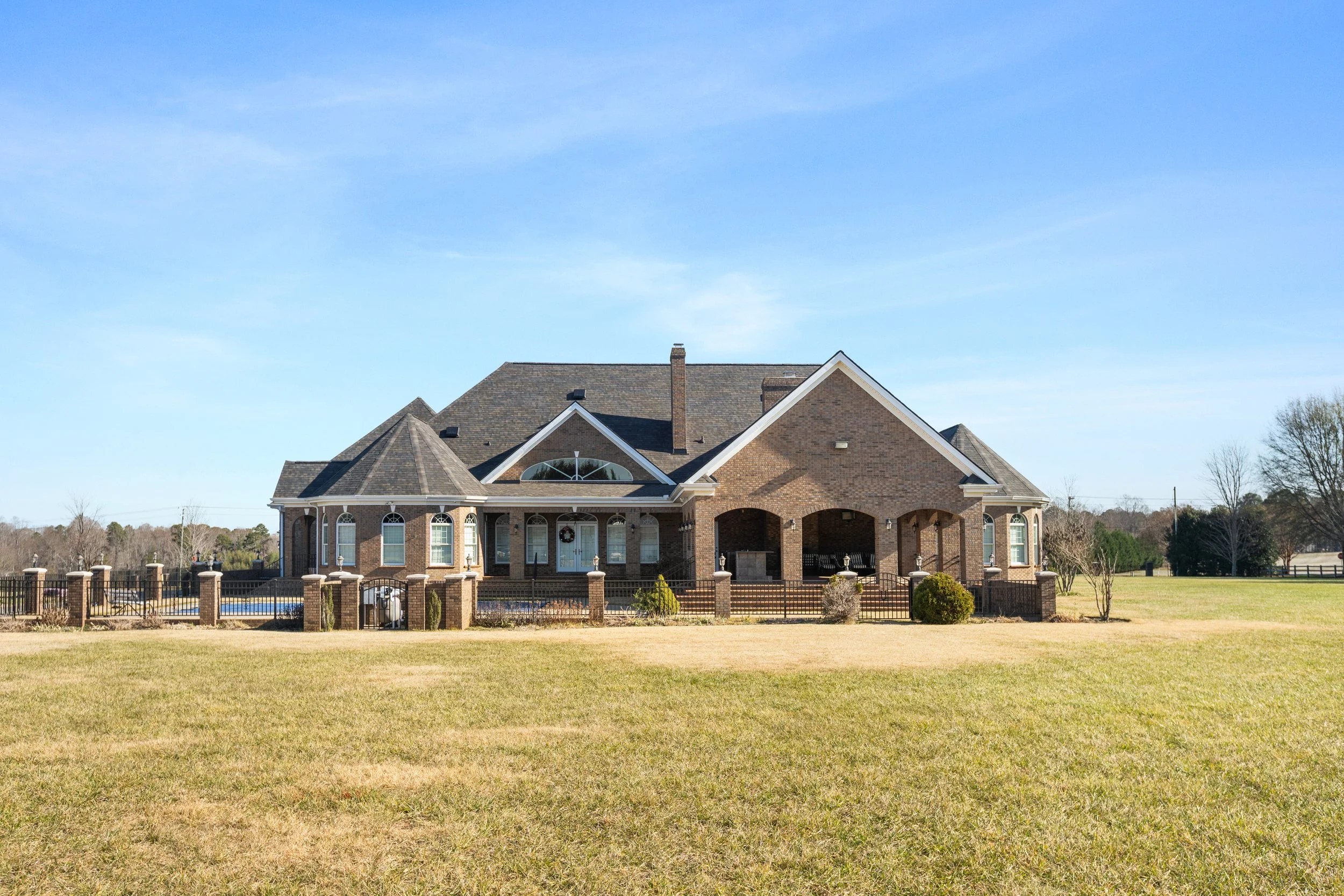 Large brick house with multiple gabled roofs, arched windows, and a covered porch, surrounded by a fenced yard with grass and landscaping, under a clear blue sky.