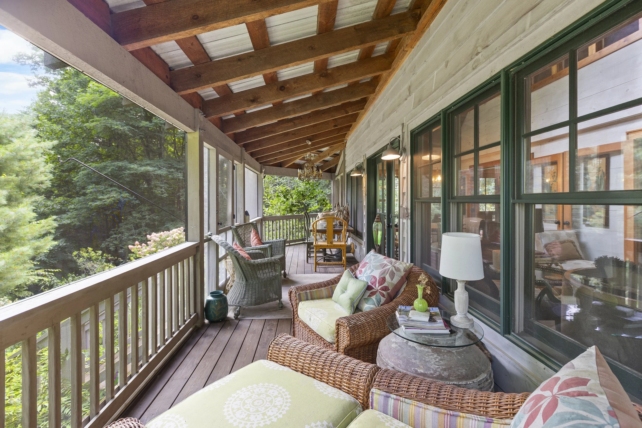 A porch with wicker and wooden furniture, cushions, a lamp, and surrounded by trees.