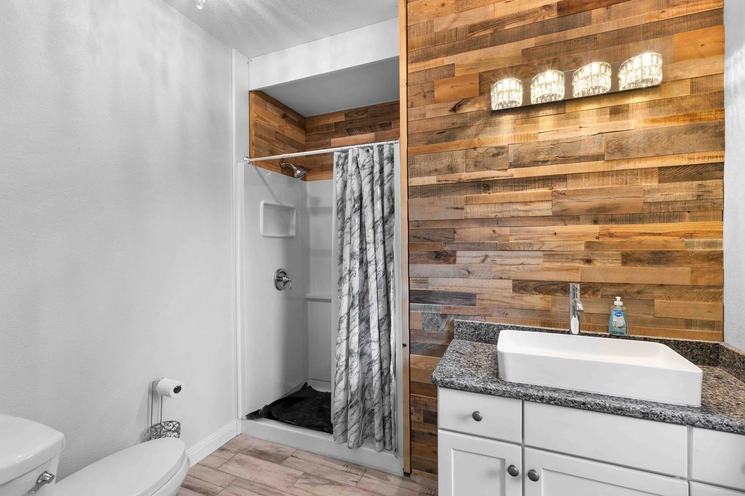 Bathroom with a white toilet, a shower with a marble-patterned curtain, a granite countertop with a white sink, and a wooden accent wall with a light fixture.