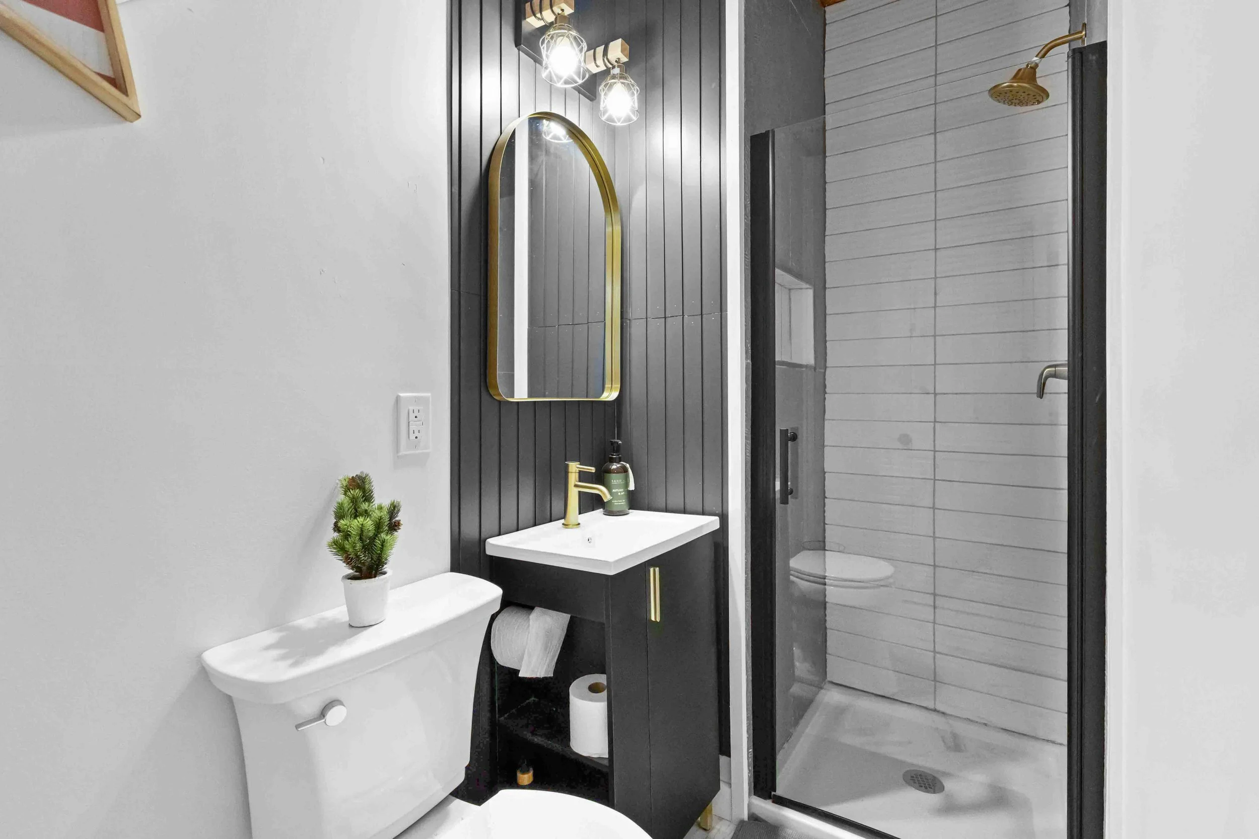 A modern bathroom featuring a black and white color scheme with gold accents. There is a small sink with a gold faucet, a mirror with an arched gold frame, a toilet with a small potted plant on top, and a shower with a black frame and a gold showerhe