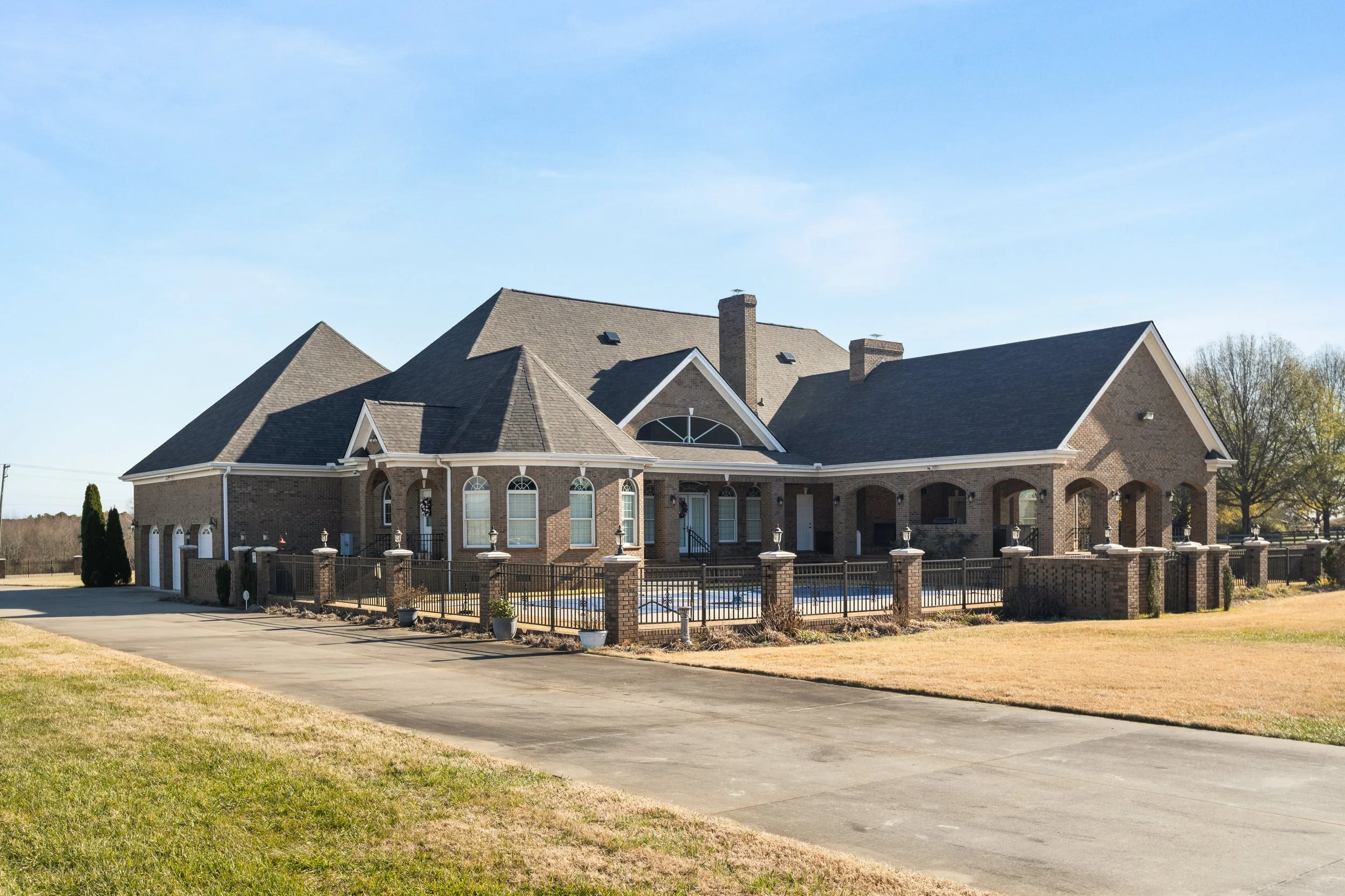 A large brick house with a gated backyard pool, surrounded by a brick fence and situated on a spacious lawn under a clear blue sky.