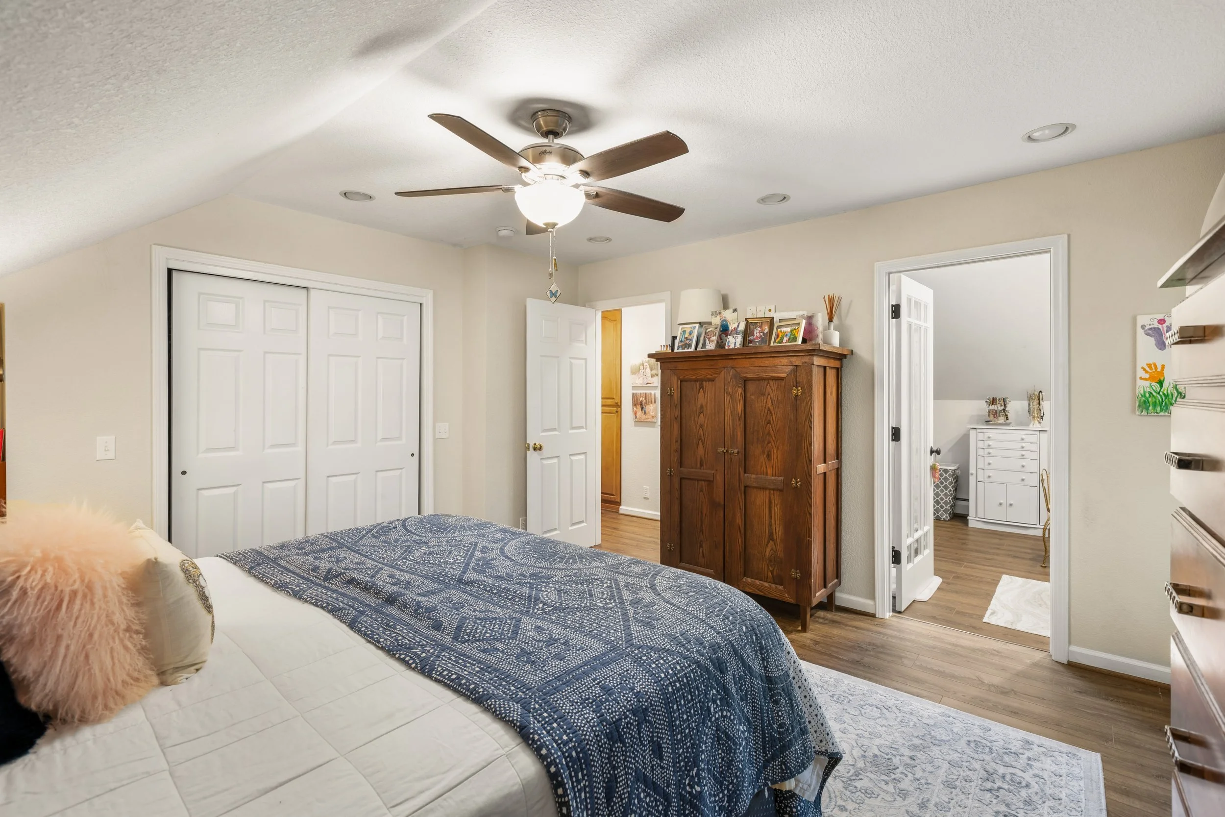 A bedroom with a bed covered in a blue patterned quilt, a furry pink pillow, and a beige pillow, wooden flooring, a wooden wardrobe with family photos on top, open closet doors, a ceiling fan, and a doorway leading to a small room with a white dresse