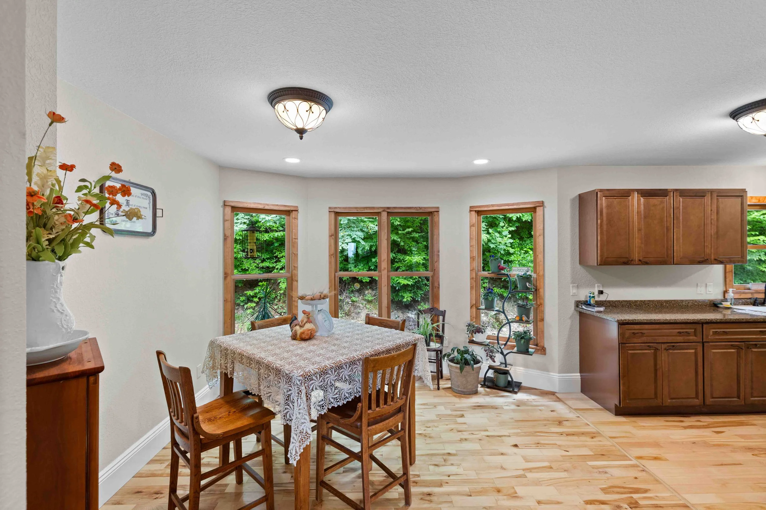 A dining area with a wooden table covered by a lace tablecloth, surrounded by four wooden chairs. The room has three windows with natural wood trim showing greenery outside. To the left, there is a white vase with orange and white flowers. On the rig