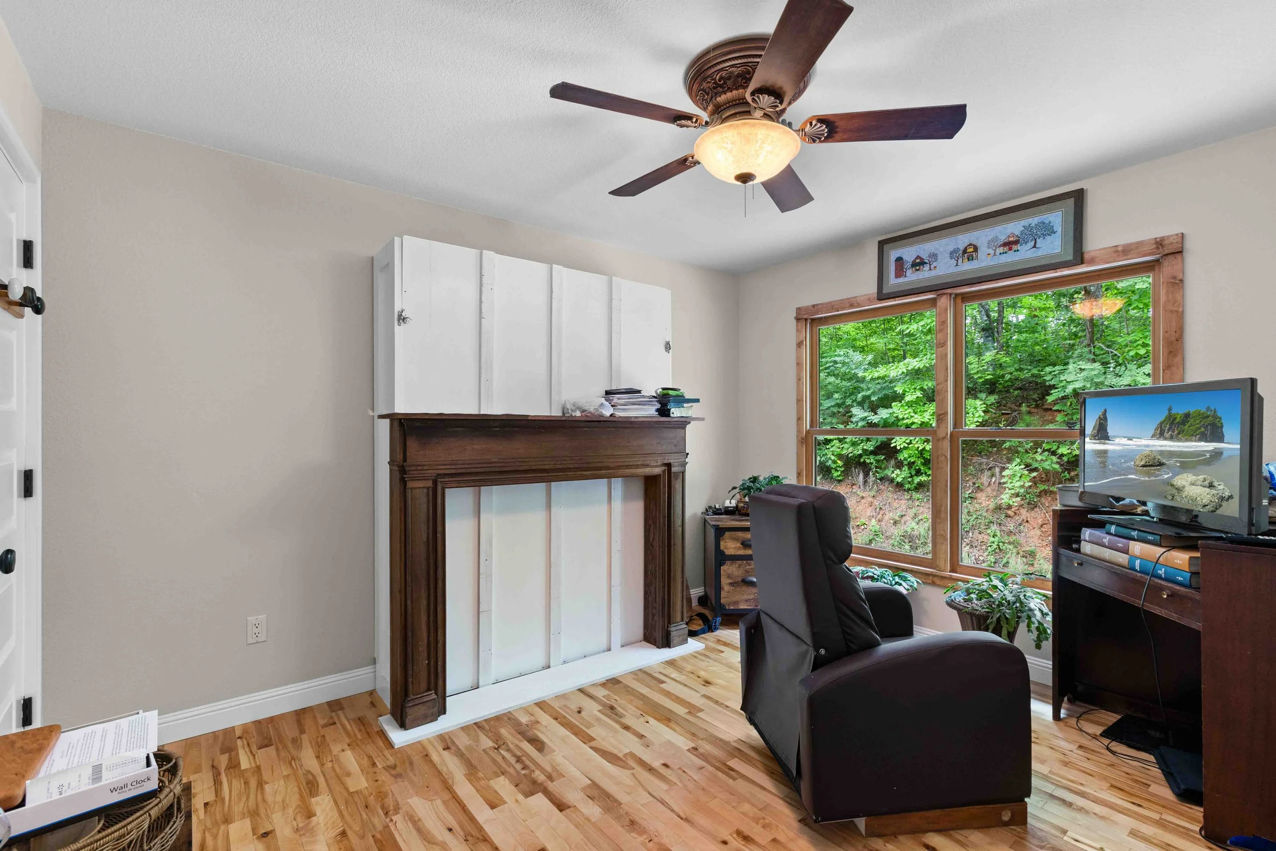 A living room with a ceiling fan, a window overlooking greenery, a black armchair, a small wooden side table, a flat-screen TV on a wooden stand, and art above the window.