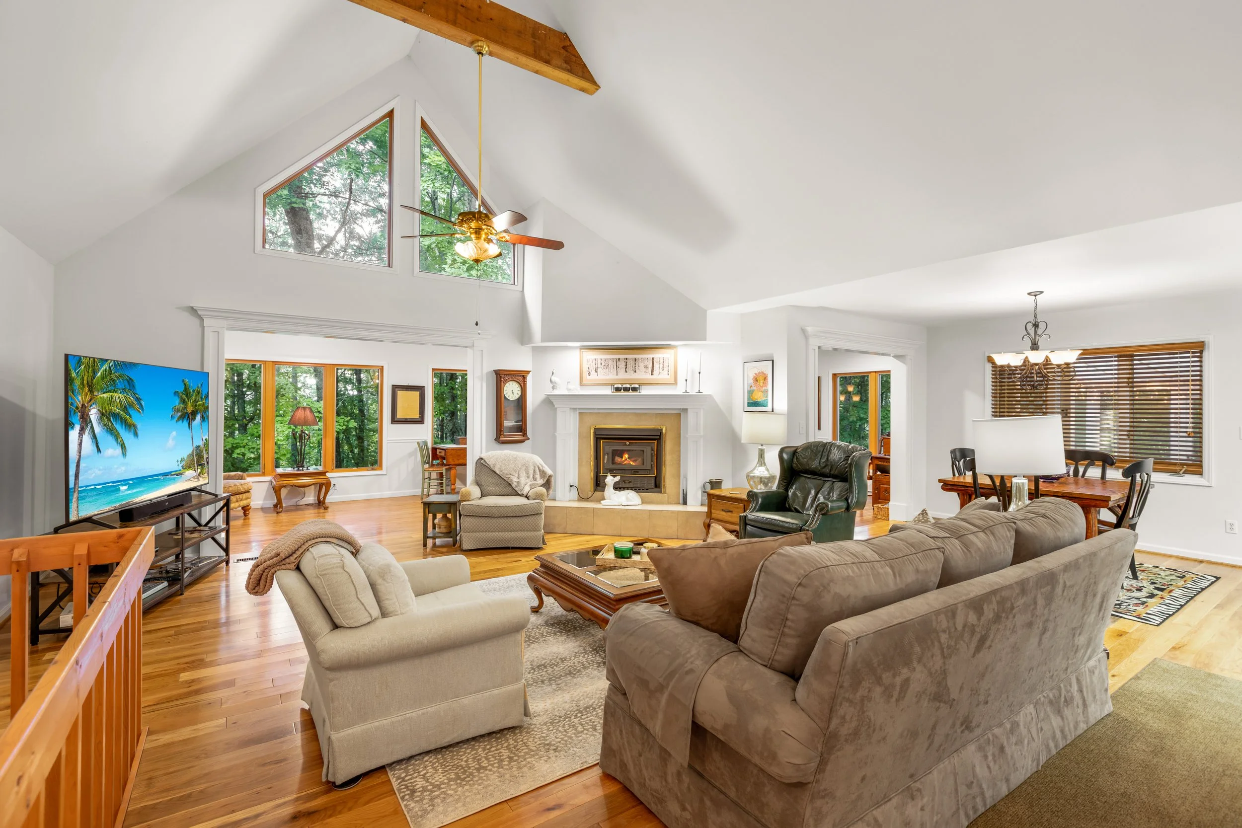 Spacious living room with high vaulted ceilings, large windows, hardwood floors, fireplace, and various seating arrangements including a sofa, armchairs, and recliner. There is a flat-screen TV on a stand, a ceiling fan, and decorative elements like 