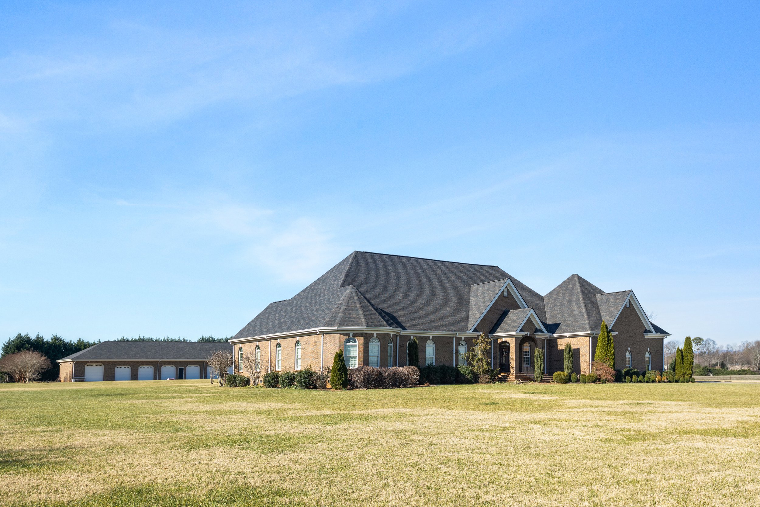 Large brick house with multiple peaked roofs, surrounded by well-maintained bushes and trees, on a grassy lawn under a clear blue sky.