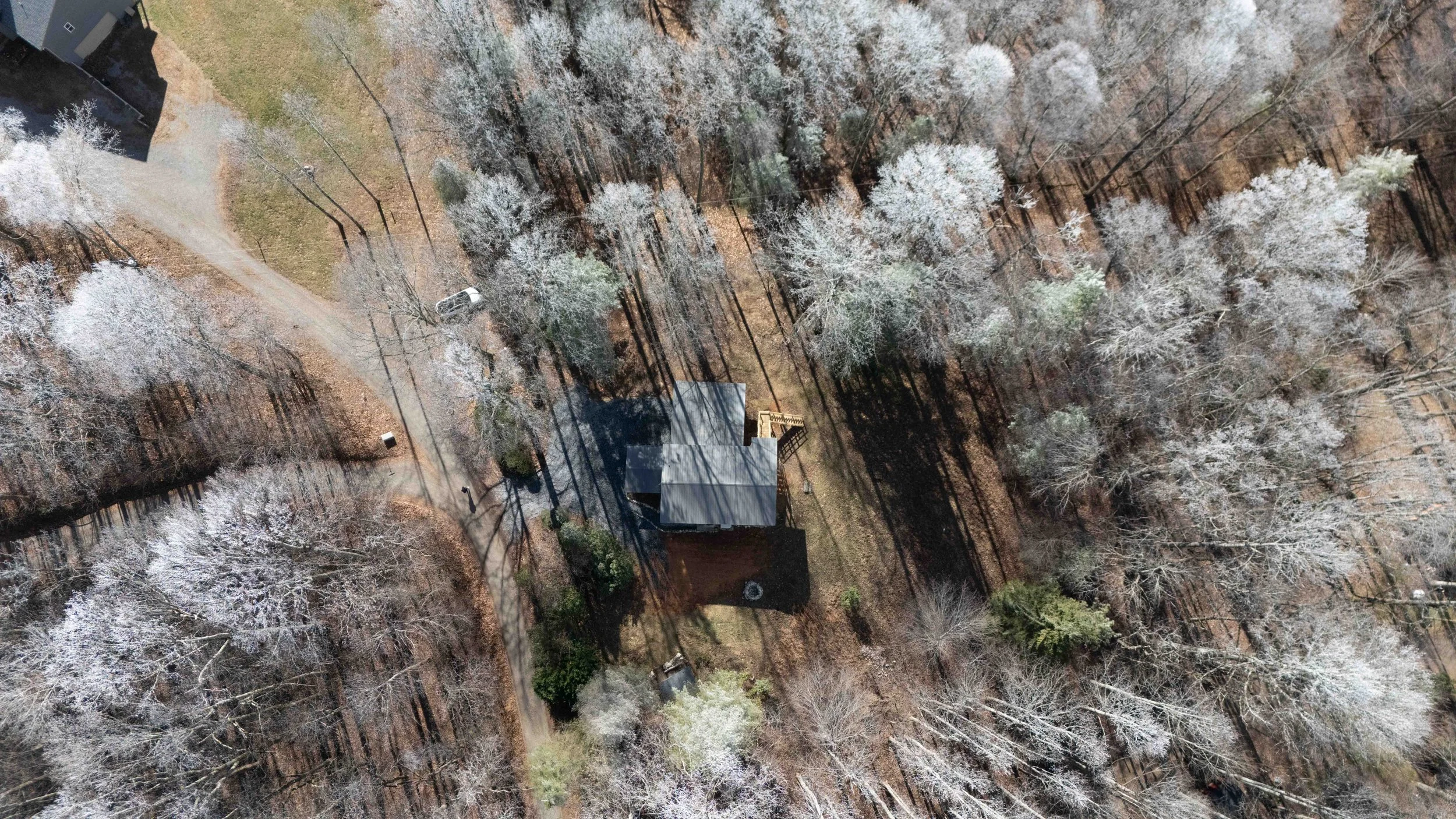 Aerial view of a house surrounded by a wooded area with bare trees, some of which have snow on their branches, and a winding driveway.