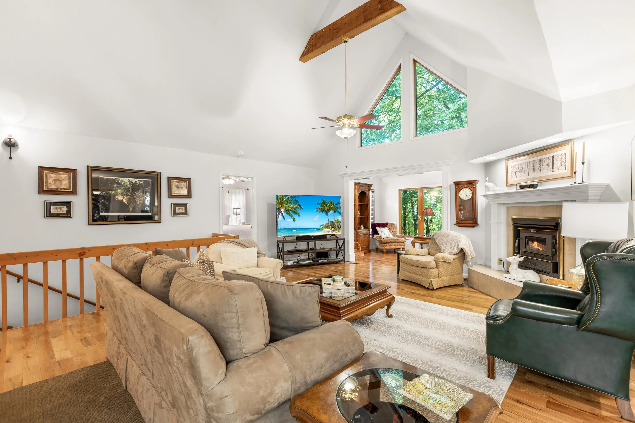 Living room with high vaulted ceiling and large triangular window showing green trees outside, beige sofa, green armchair, fireplace, wooden furniture, and a television displaying a beach scene.