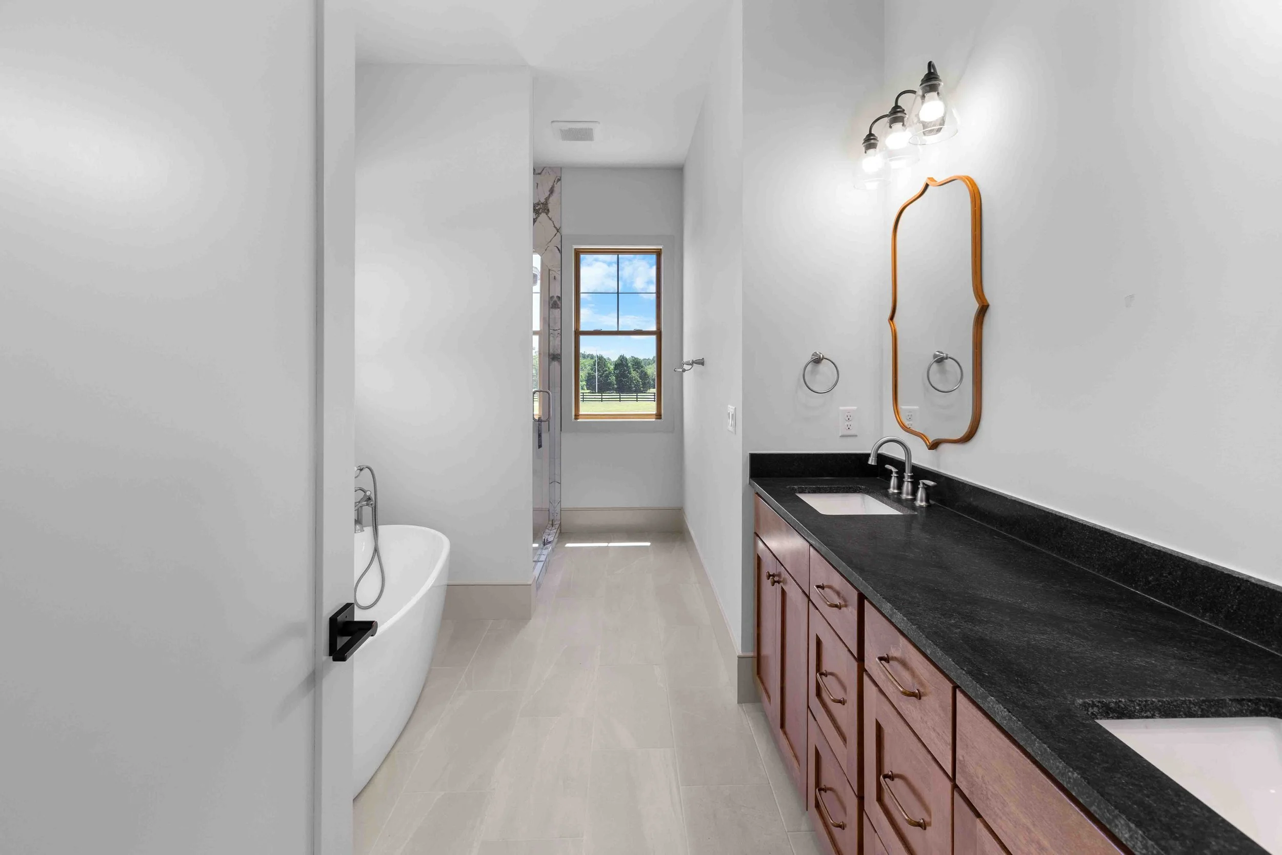 Modern bathroom with a black countertop vanity, two sinks, wooden cabinets, a large mirror, and three light fixtures, with a bathtub, a window showing outdoor scenery, and a walk-in shower with glass door.
