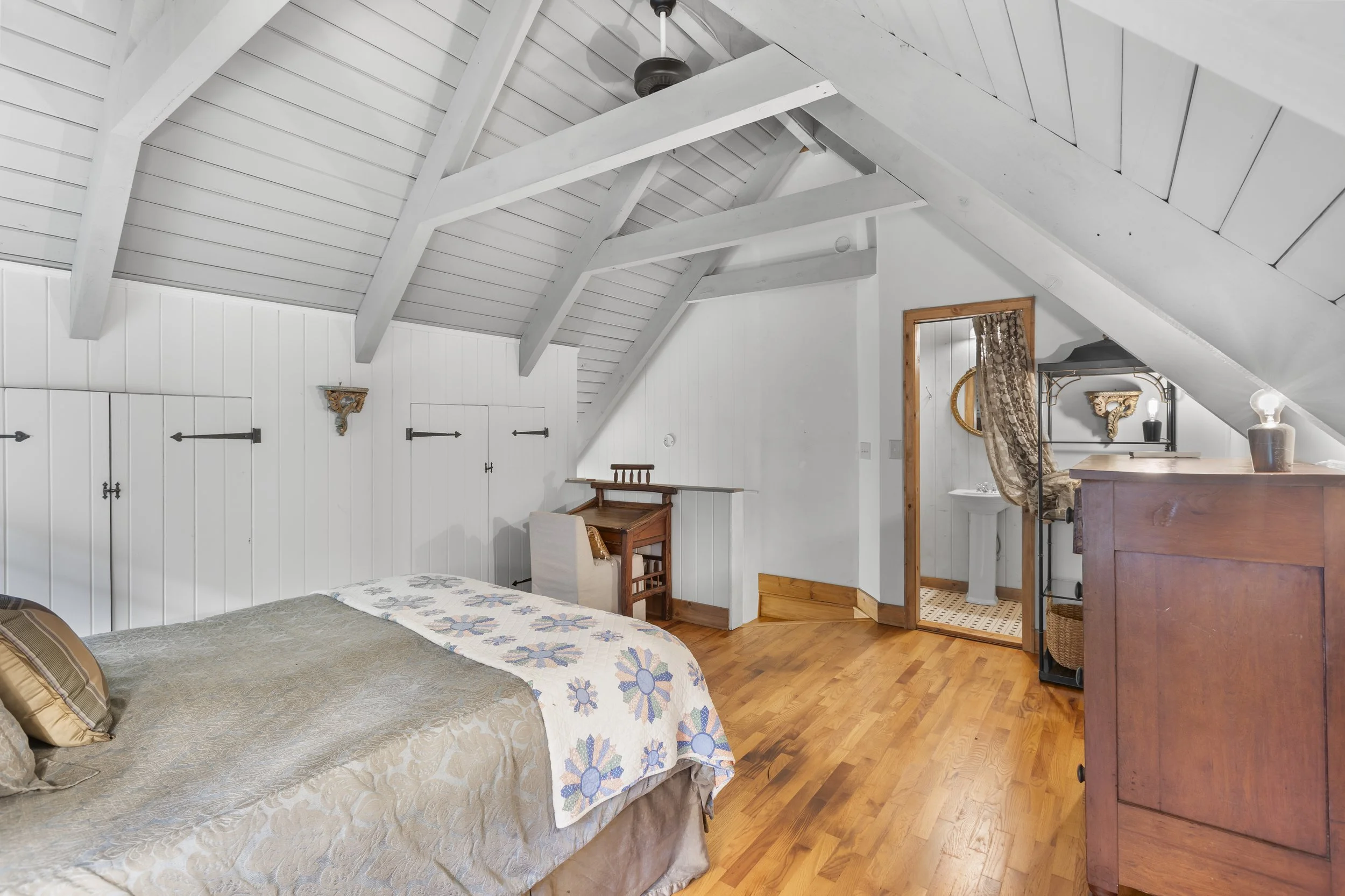 A bedroom with a sloped ceiling and white wooden beams, containing a bed with beige embroidery, a small desk, and a wooden dresser. An open door shows a bathroom with a pedestal sink and patterned tile floor.