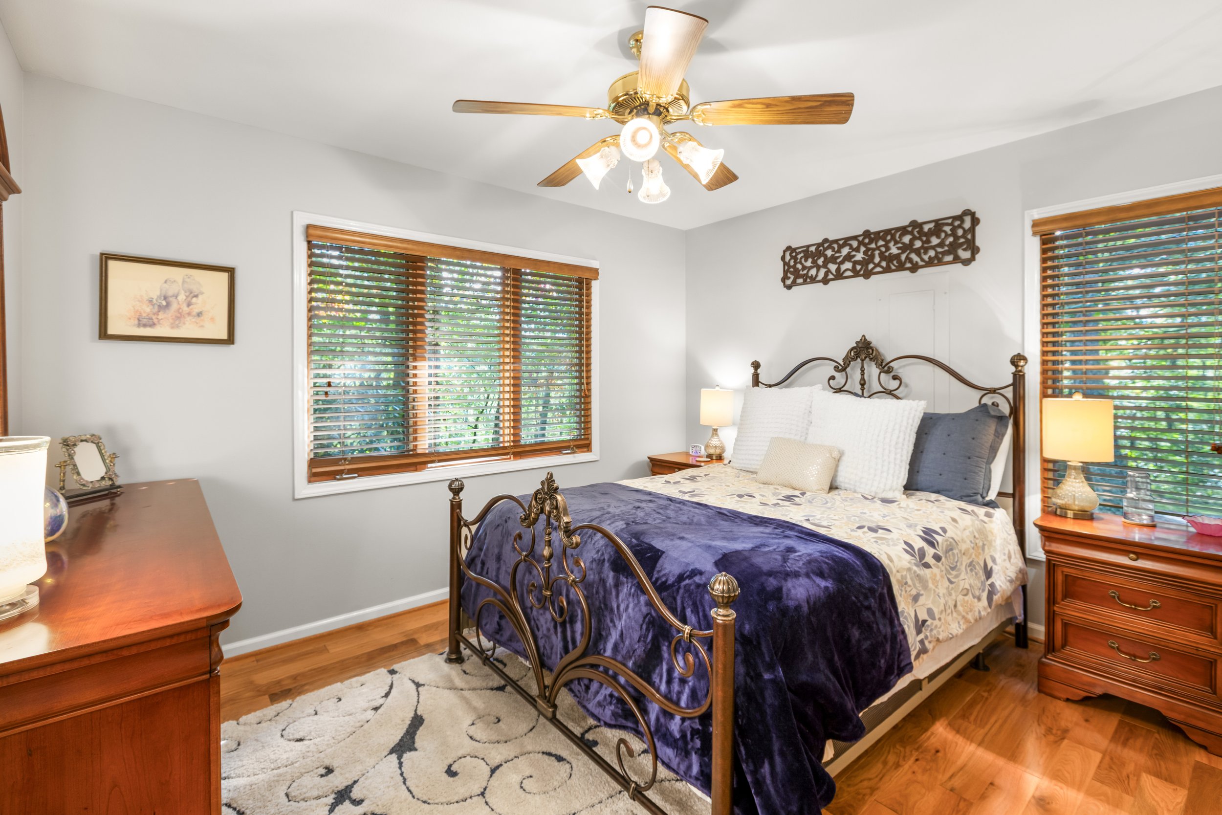 A bedroom with a metal bed frame, wooden nightstands, and light-colored walls. There are two lamps on each nightstand, and windows with wooden blinds on two sides of the room. The bed is made with a floral comforter, pillows, and a purple throw blank