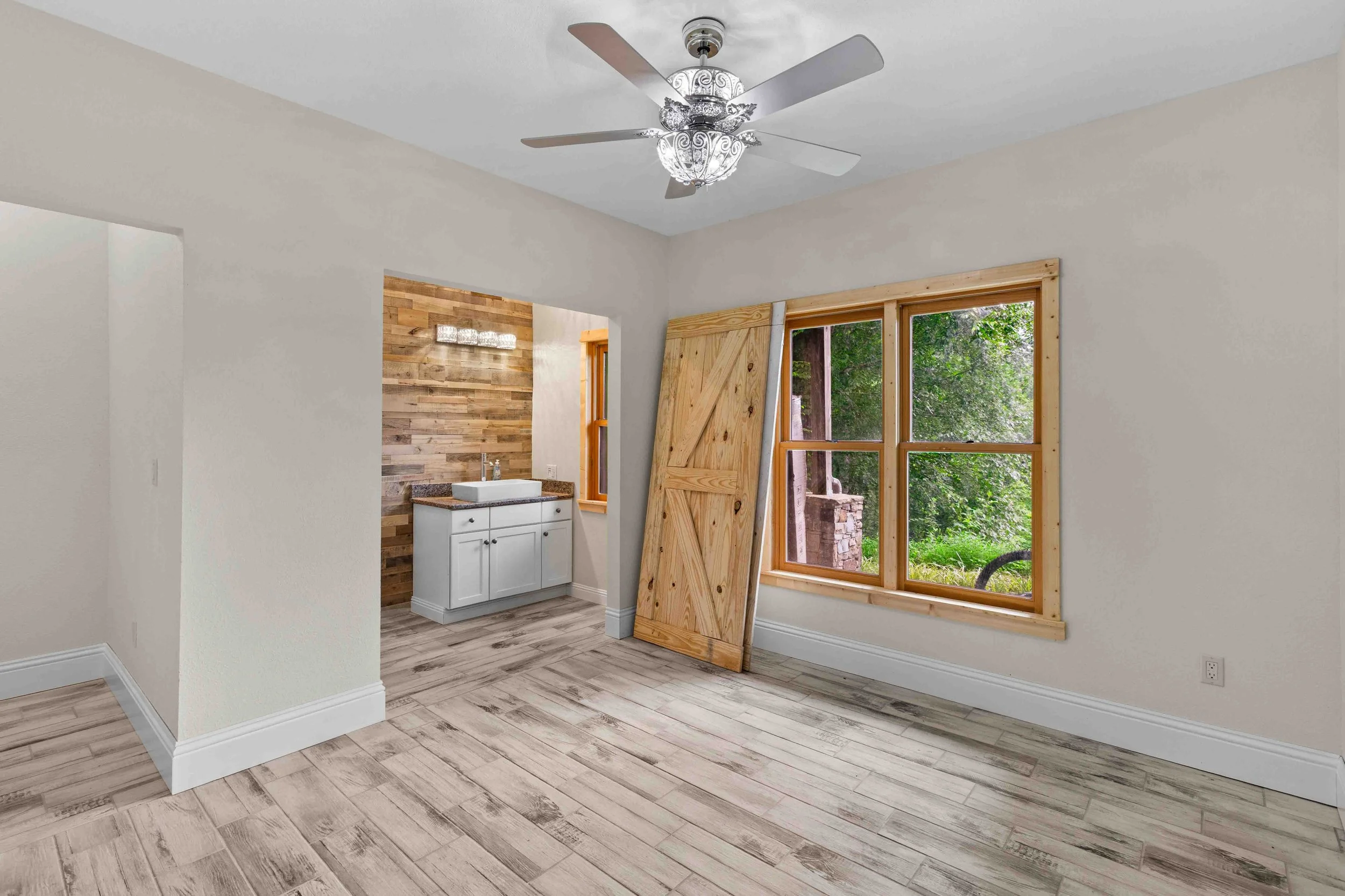 Empty room with light-colored walls, wood flooring, large window with natural scenery, ceiling fan, and a small attached bathroom with wooden accent wall and white sink cabinet.