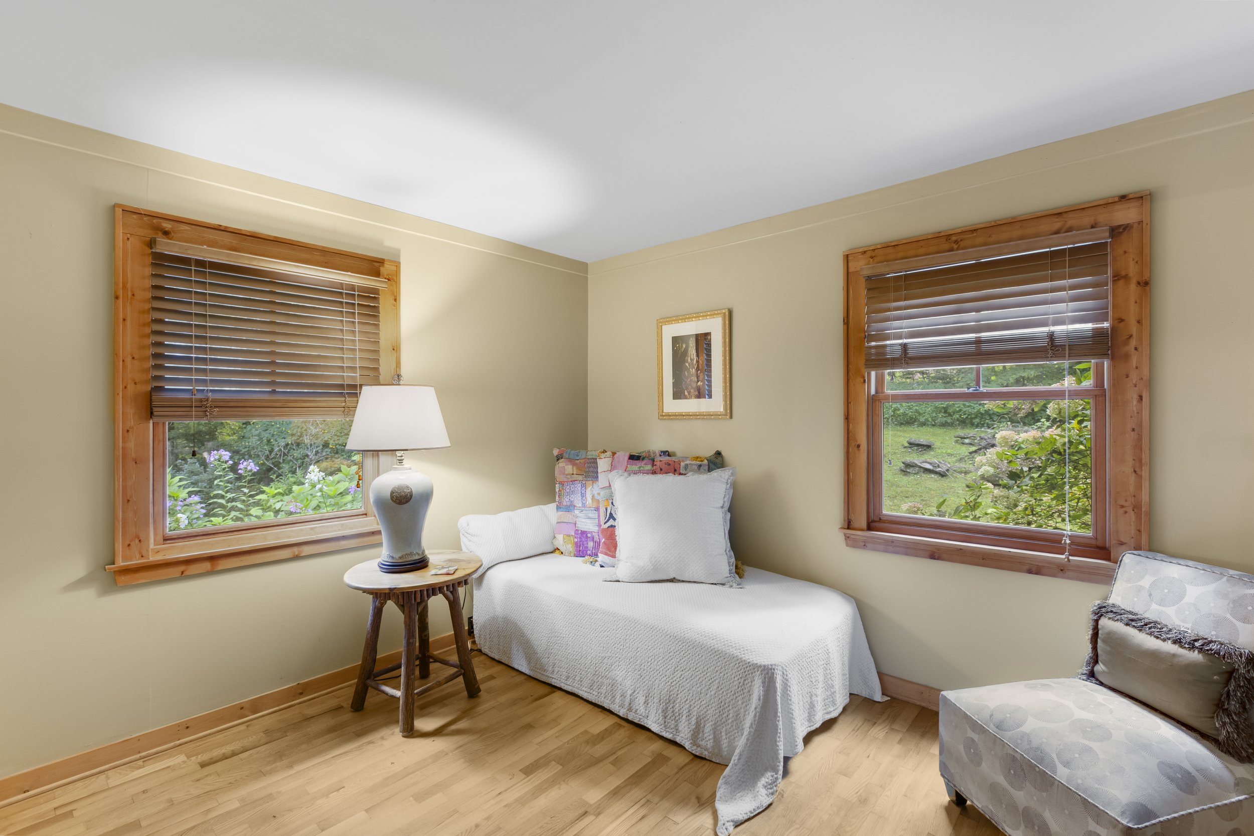 A cozy bedroom with two windows featuring wooden frames and blinds, a bed with white bedding and colorful pillows, a side table with a lamp, and a patterned chair, with a framed picture on the wall.