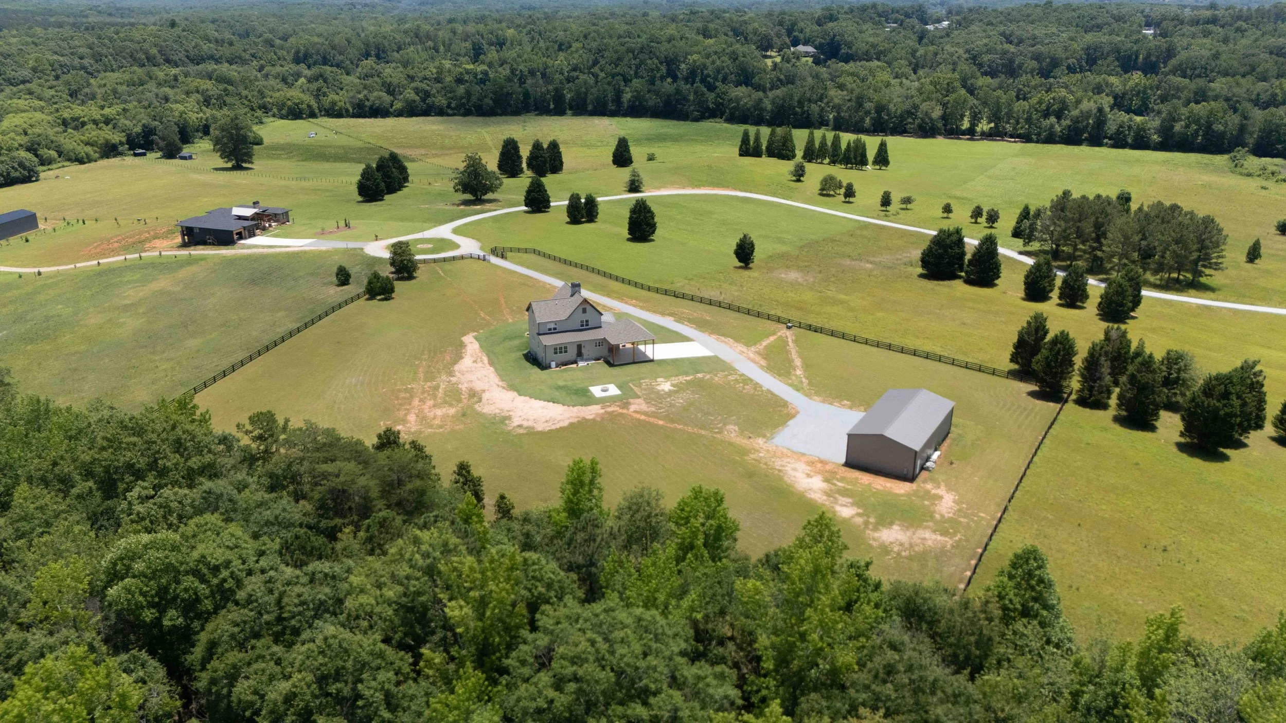 Aerial view of a rural property with a house, large lawn, trees, a barn, and a fenced area, surrounded by forest.