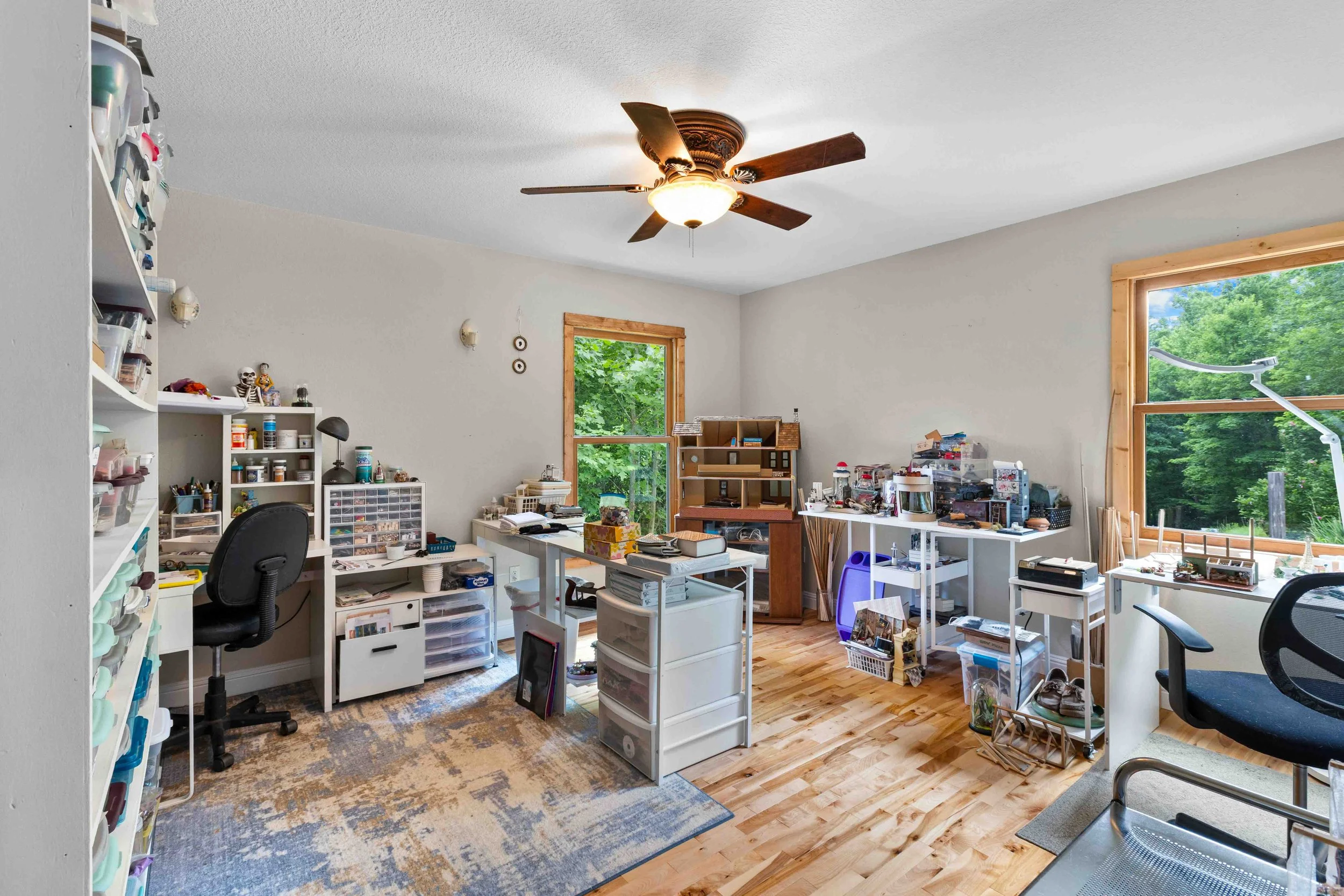 Cluttered craft room with white shelves, desks, art supplies, and large windows showing green trees outside.