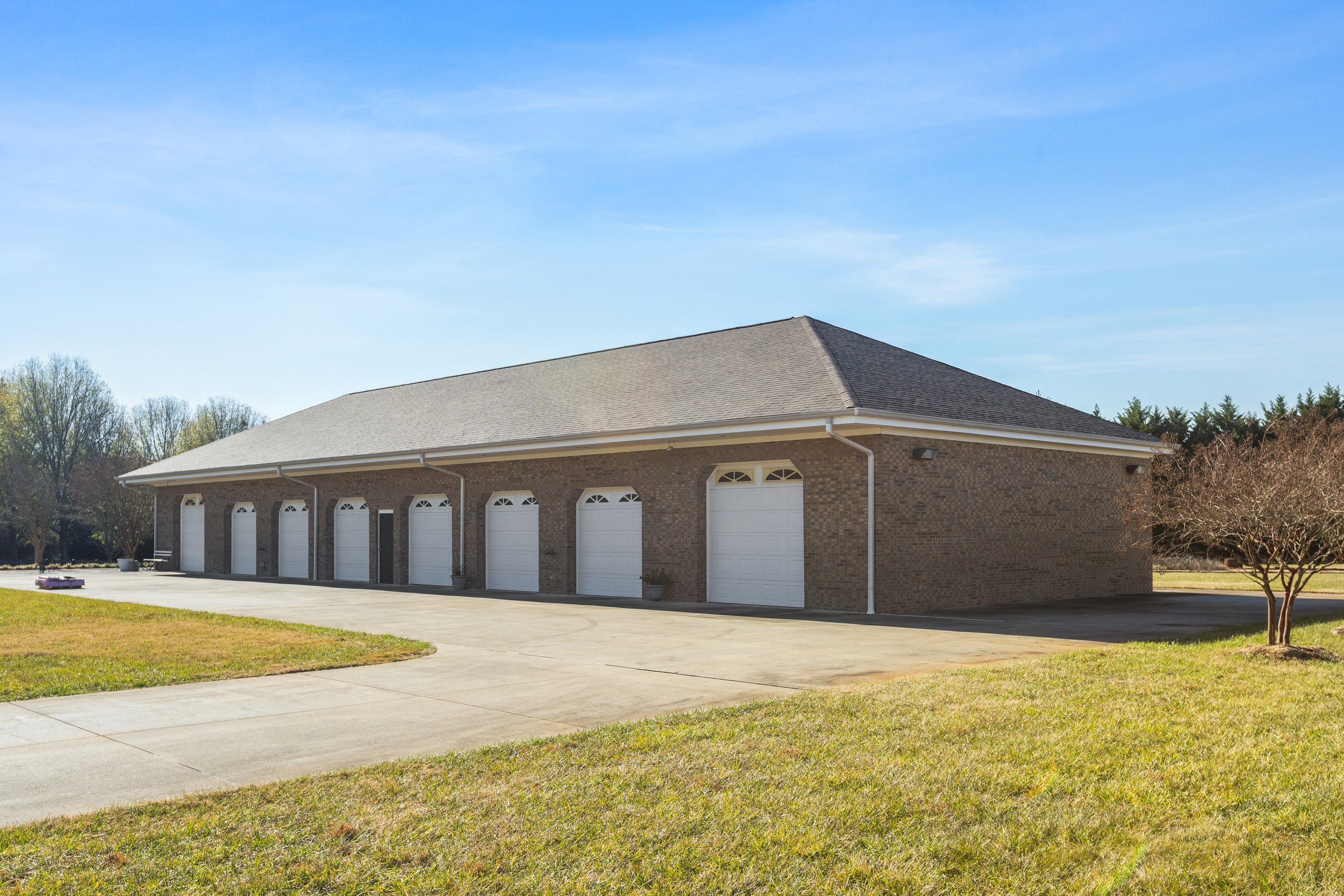 Large brick building with multiple white garage doors, a driveway in front, and surrounding trees under a blue sky.