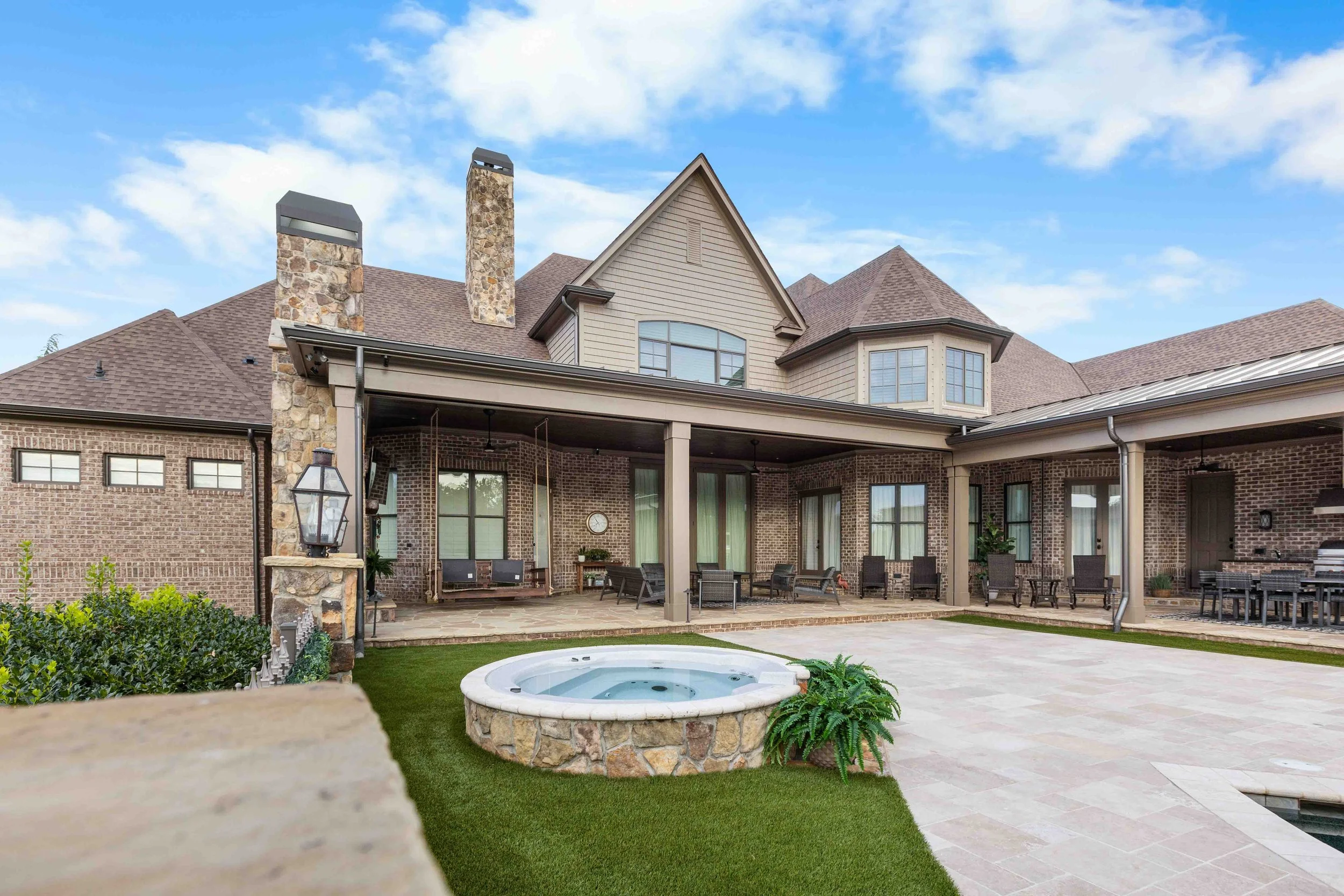 Spacious backyard of a brick house with a covered patio, outdoor furniture, a stone hot tub, and a manicured lawn under a partly cloudy sky.
