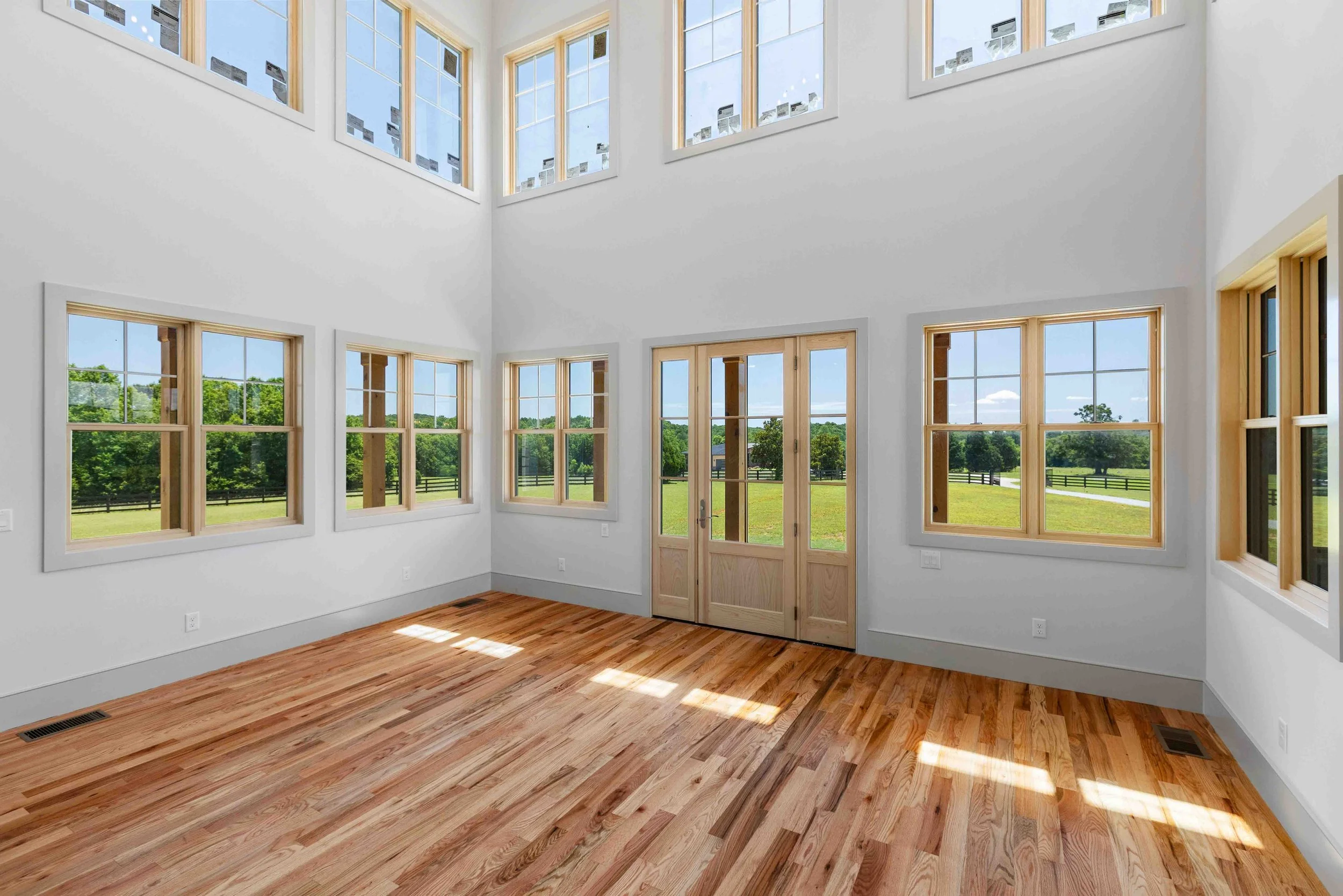 Empty room with multiple large windows, wooden floors, and a set of double wooden doors, with a view of green fields outside.