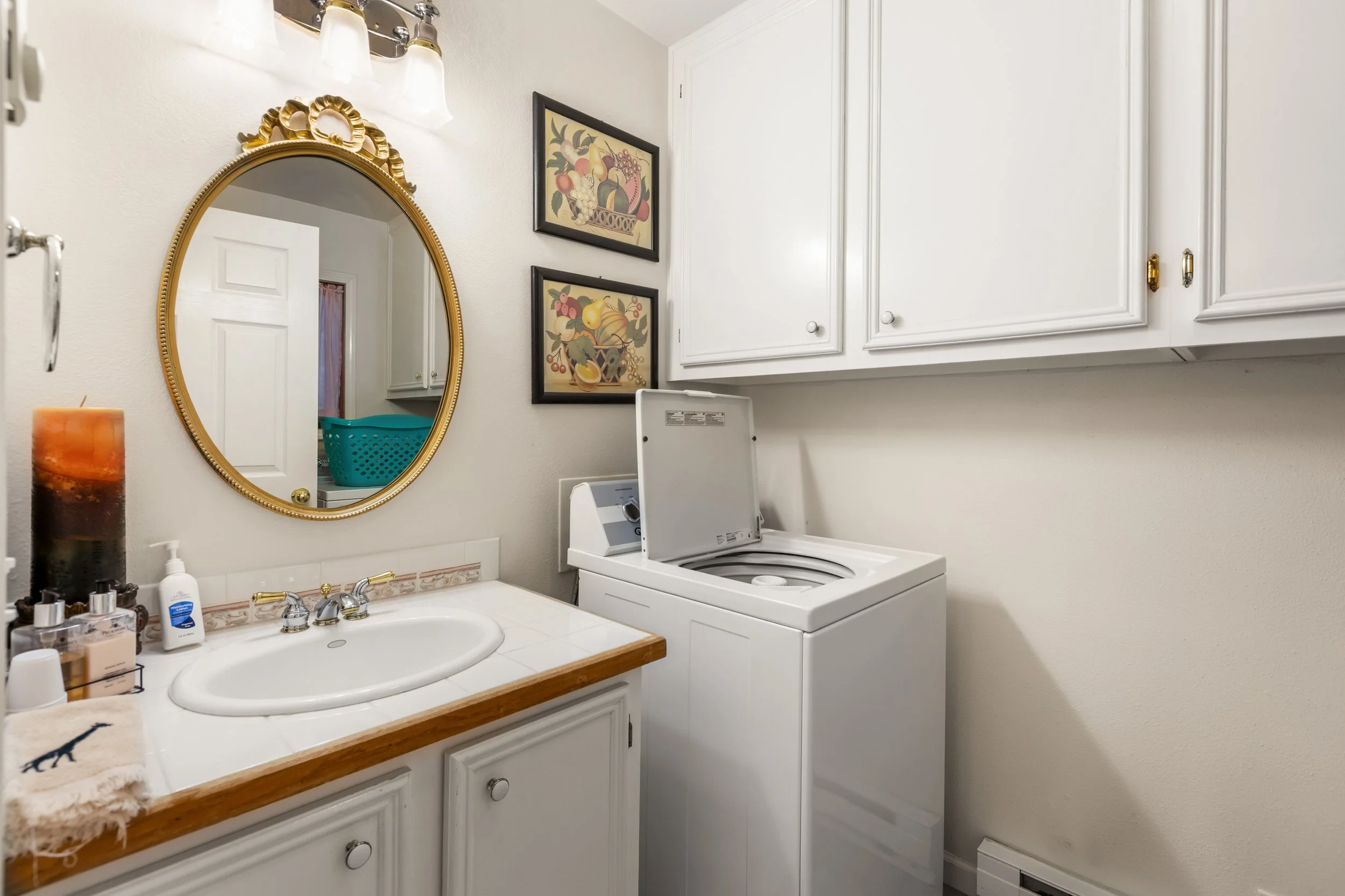 Laundry room with a washing machine, white cabinets, a bathroom vanity with a sink, wall-mounted mirror, framed fruit artwork, a candle, soap, and other toiletries.