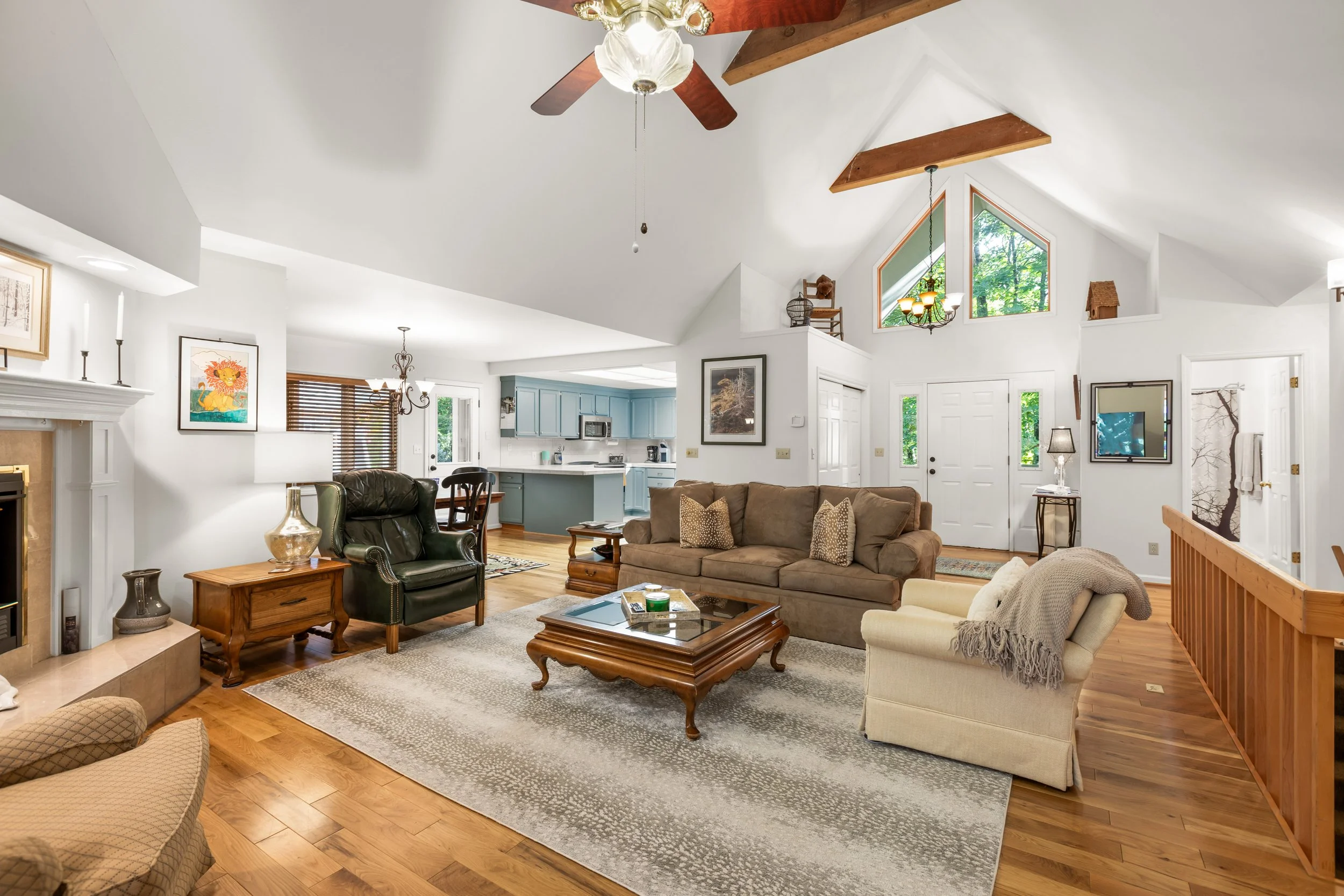 Living room with a brown sofa, beige armchair, black leather armchair, and wooden coffee table on a large gray and white rug. Fireplace with a white mantel, framed artworks on the white walls. Open kitchen with light blue cabinets and wooden flooring