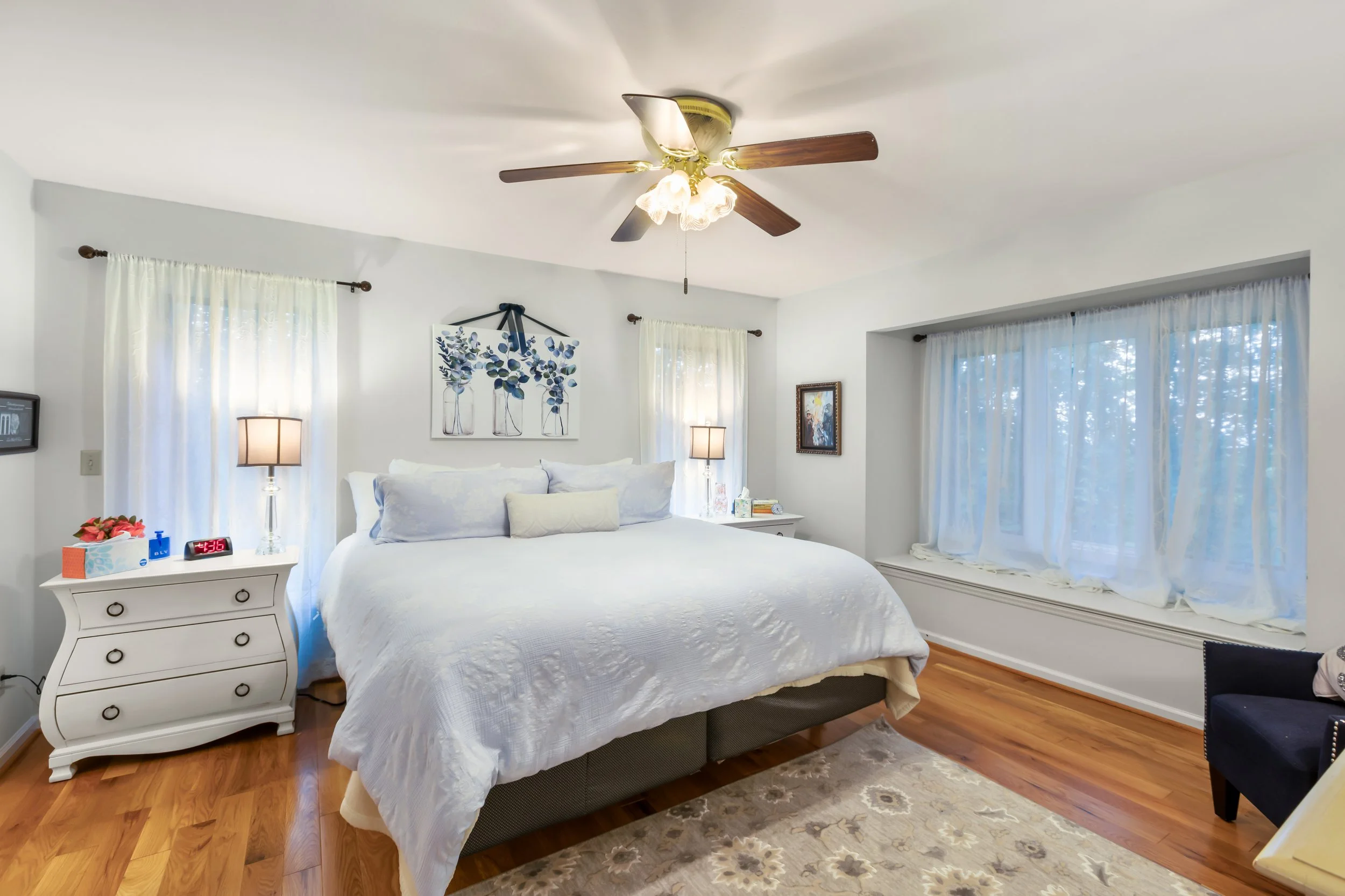 A bright bedroom with white walls, a ceiling fan with lights, a large bed with white bedding, and two windows with sheer curtains. There are nightstands with lamps on each side of the bed, a small chair to the right, and a window seat underneath the 