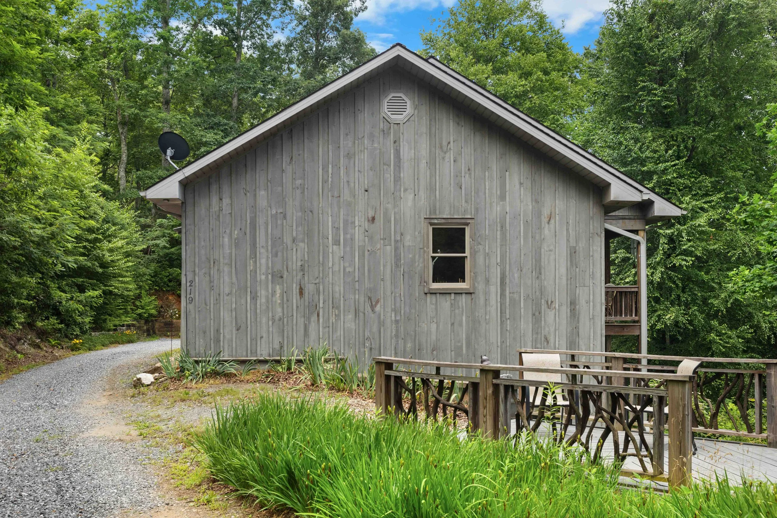 A gray wooden house with a small balcony, surrounded by greenery and trees, with a gravel pathway leading around it.