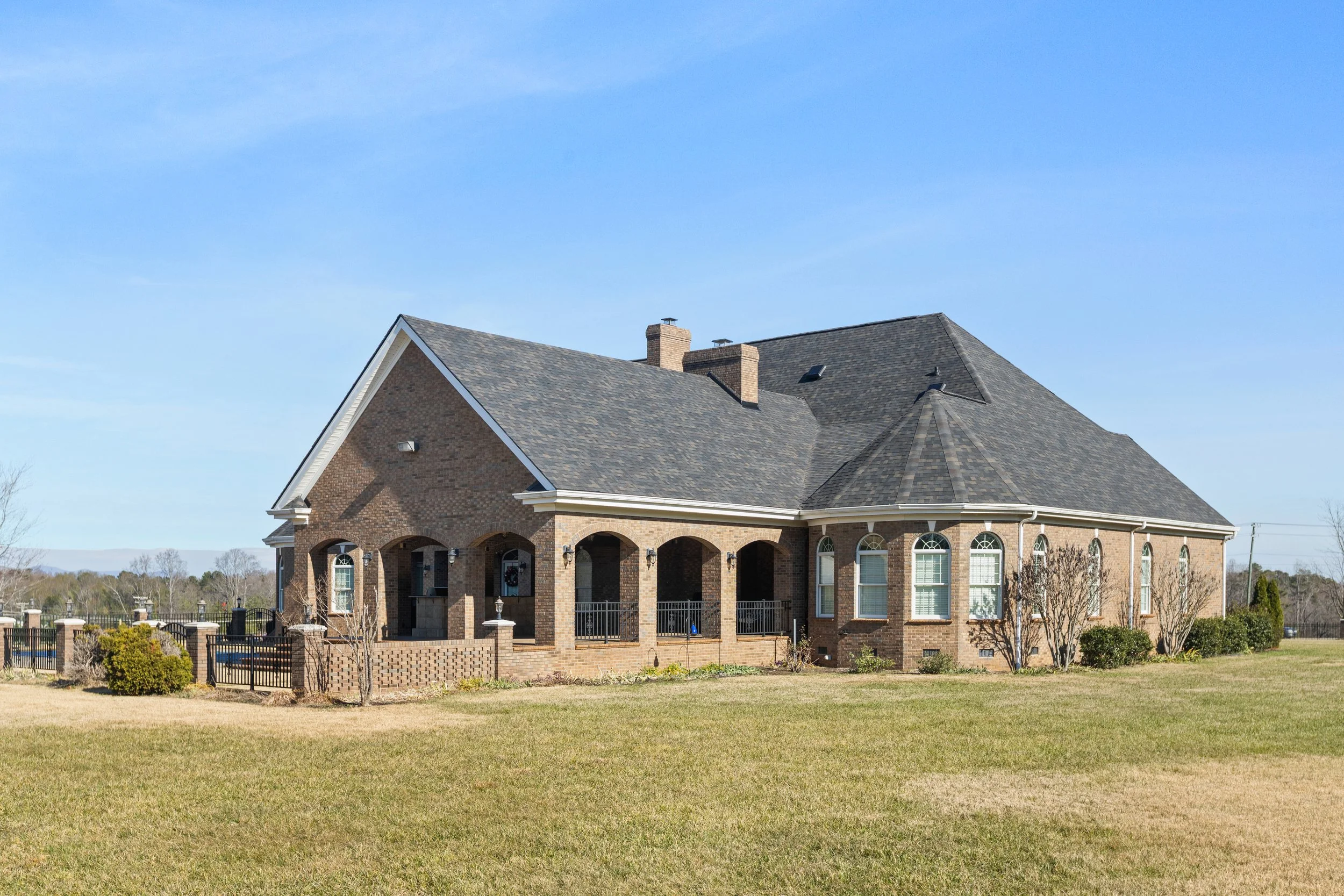 Large brick house with a gray shingle roof, white trim, arched windows, and a screened porch, surrounded by a grassy yard and a black fence under a clear blue sky.
