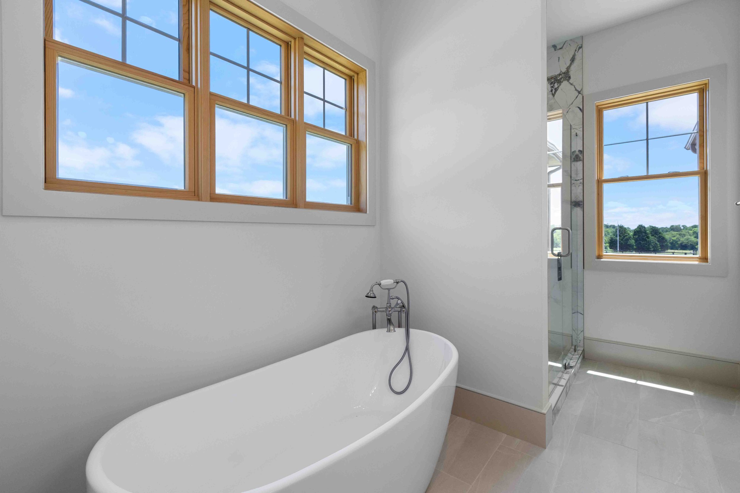 A modern bathroom with a freestanding bathtub below a large window with wooden frames, and a walk-in shower with a glass door and window showing a scenic outdoor view.