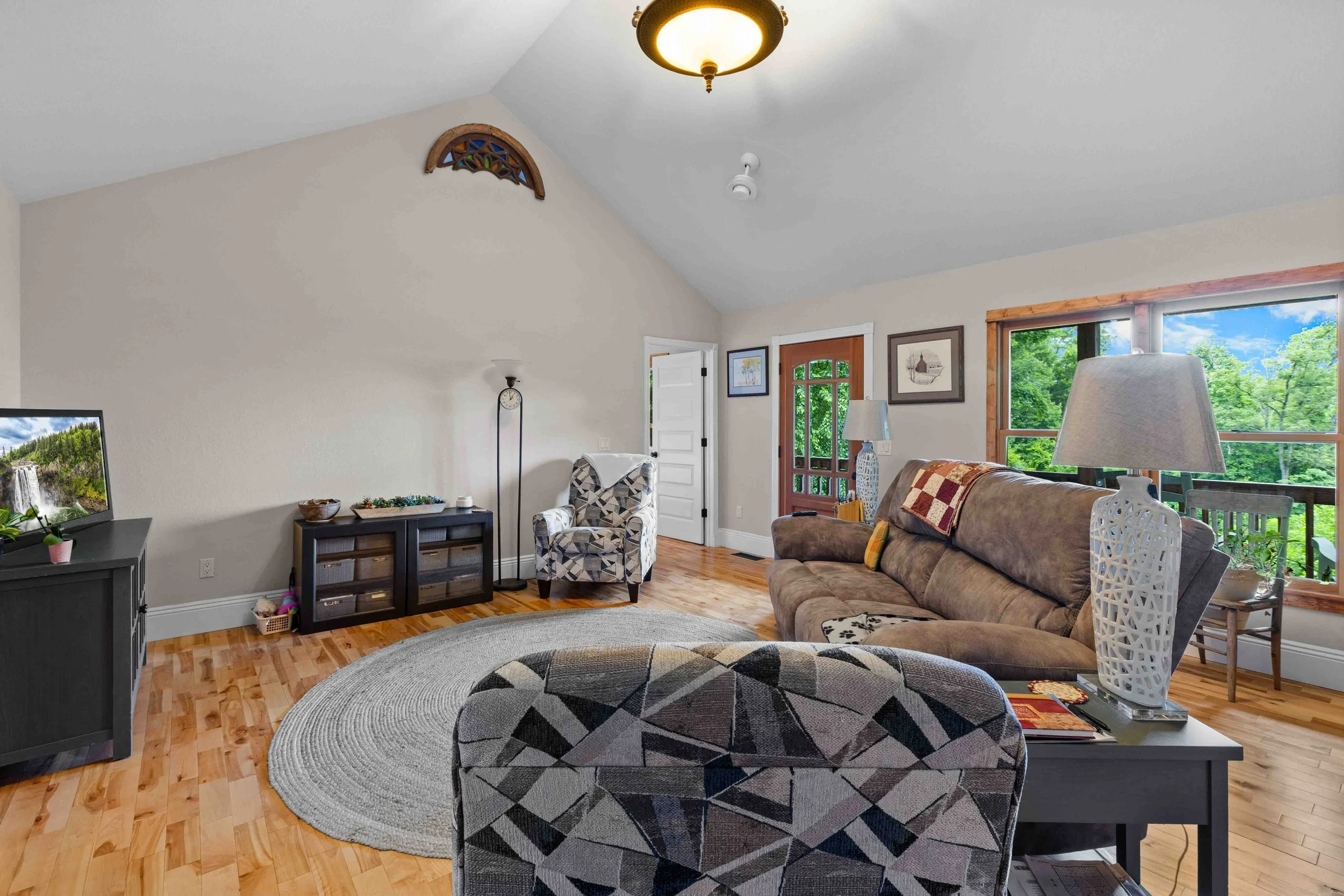 Living room with a brown couch, patterned armchair, black TV stand, flat-screen TV, white walls, hardwood floor, large window with green trees outside, and decorative lamps on end tables.