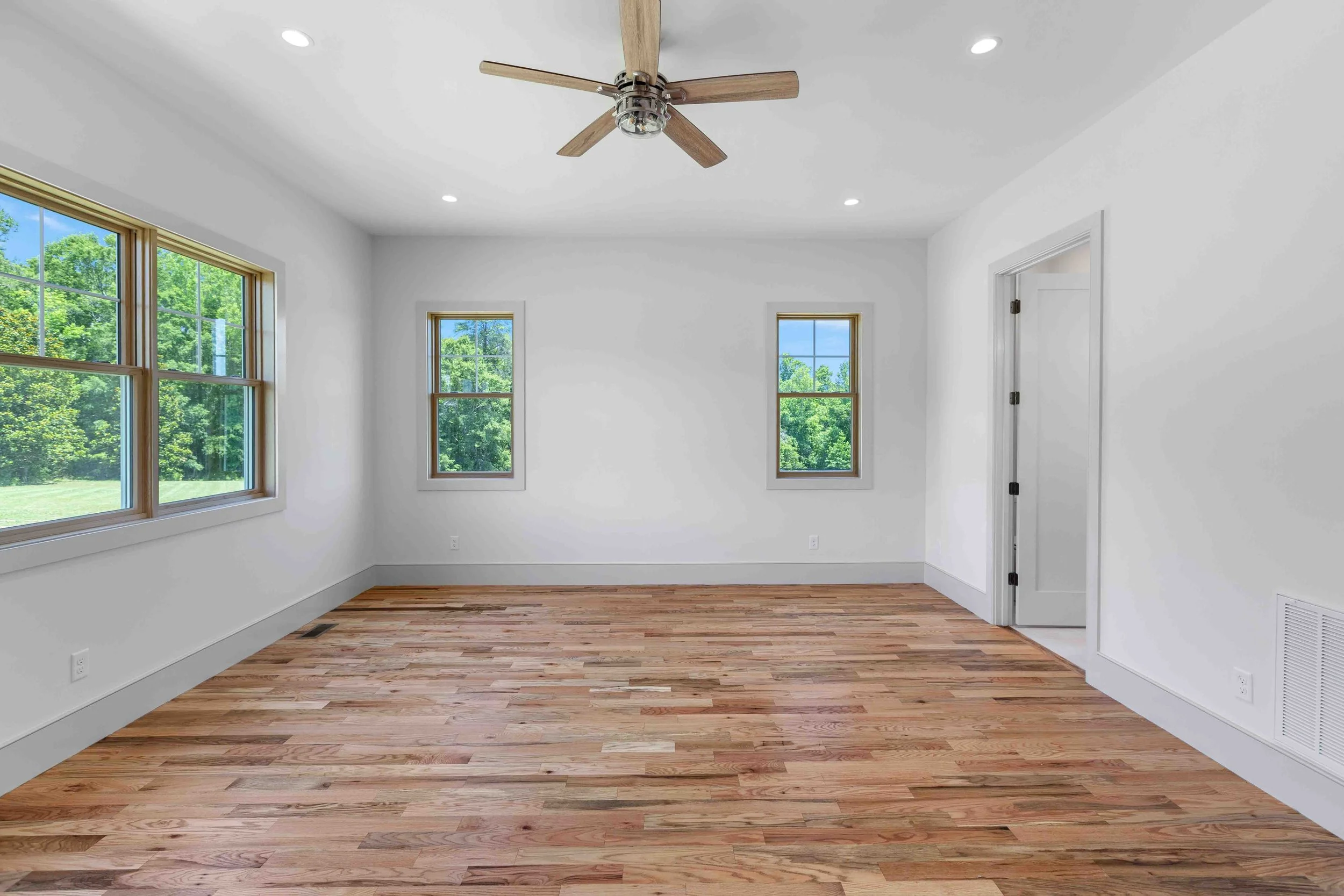 Empty room with white walls, three windows showing green trees outside, hardwood floor, ceiling fan with wooden blades, and a white door.