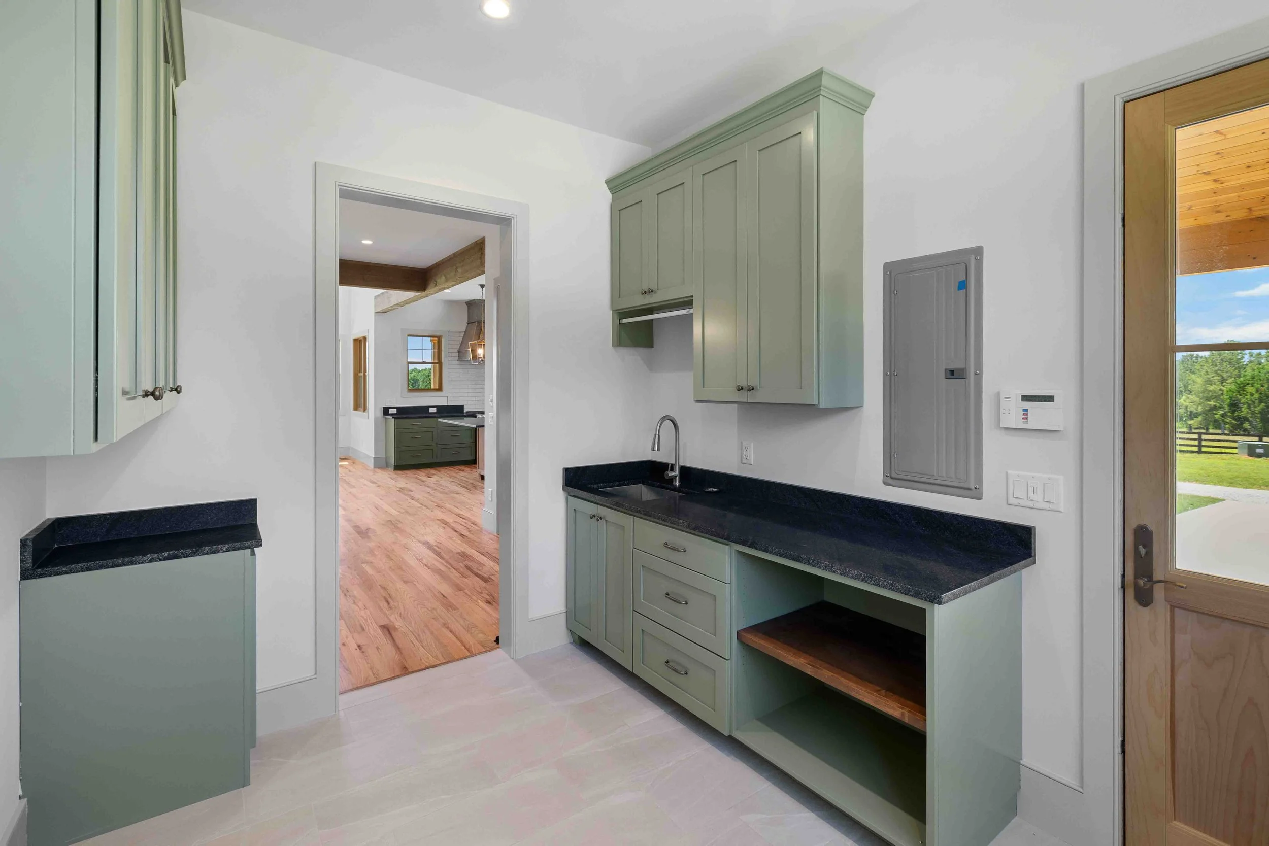 Interior view of a kitchen with green cabinetry, black countertops, and a black sink. A doorway leads to a bright room with wooden floors and green cabinets, with windows allowing natural light.
