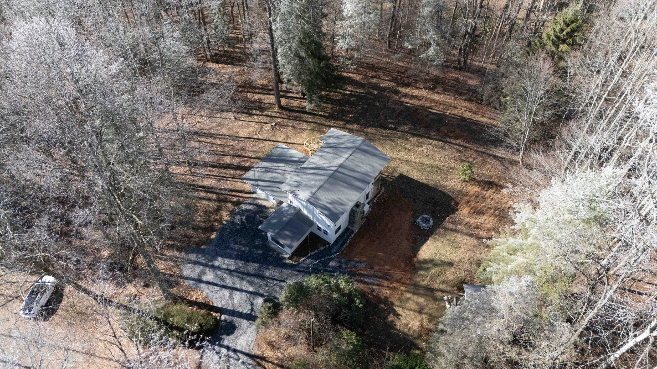 An aerial view of a house in a wooded area with leafless trees and a driveway with a parked vehicle.