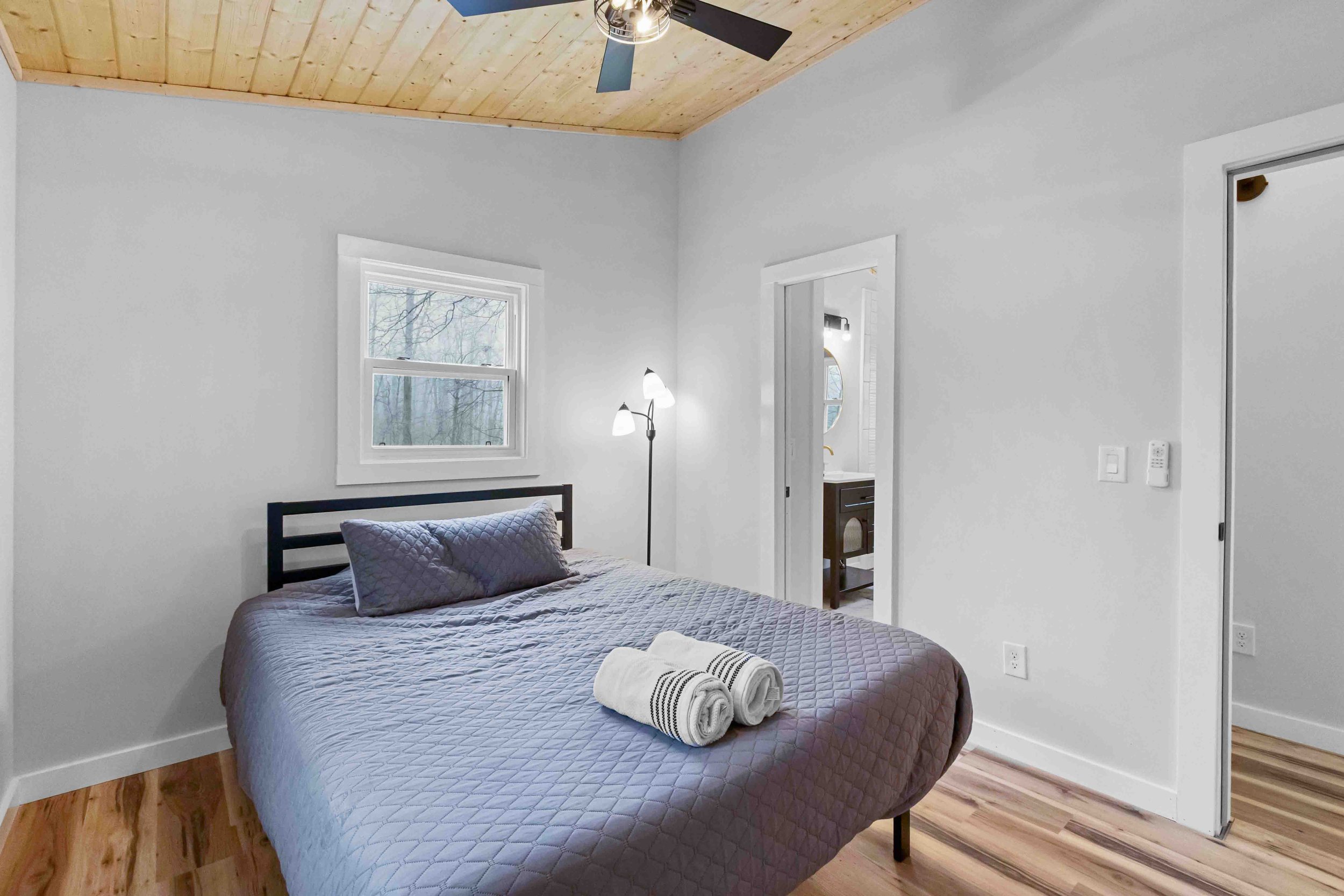 A bedroom with a bed covered in a gray quilt and matching pillows, two rolled white towels with black stripes, a window showing trees outside, a floor lamp beside the bed, and an attached bathroom visible through an open door, featuring a sink with a