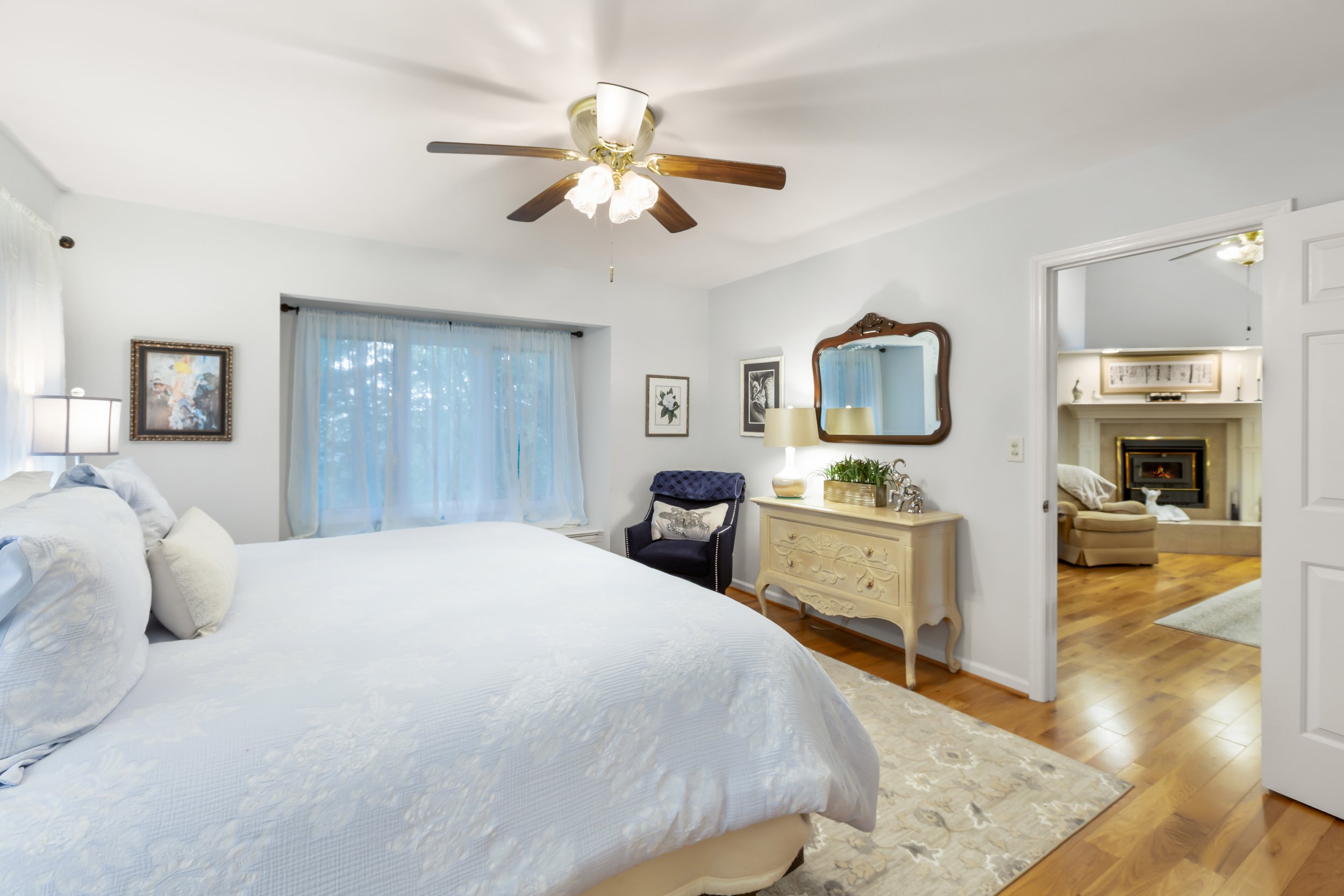 Cozy bedroom with white walls, a white bed, a window with sheers, a ceiling fan, framed artwork, a dark armchair with a pillow, a cream-colored dresser with a mirror, and a view into a living room with a fireplace.