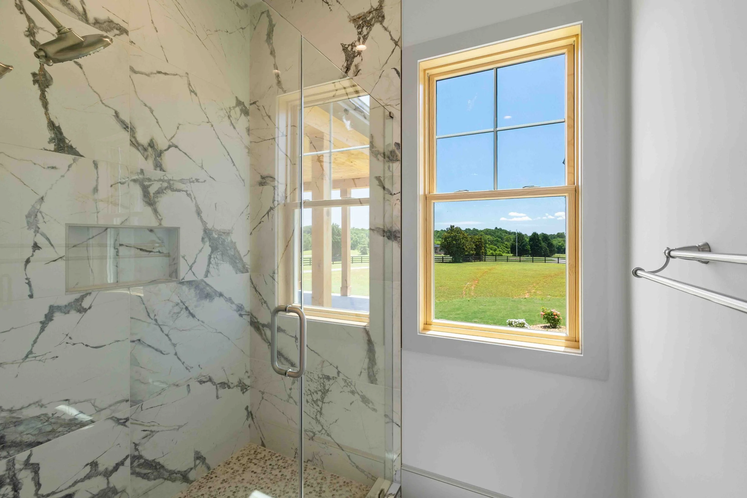 The image shows a modern shower with marble walls and a glass door, next to a window with a wooden frame offering a view of a grassy field and trees under a blue sky.