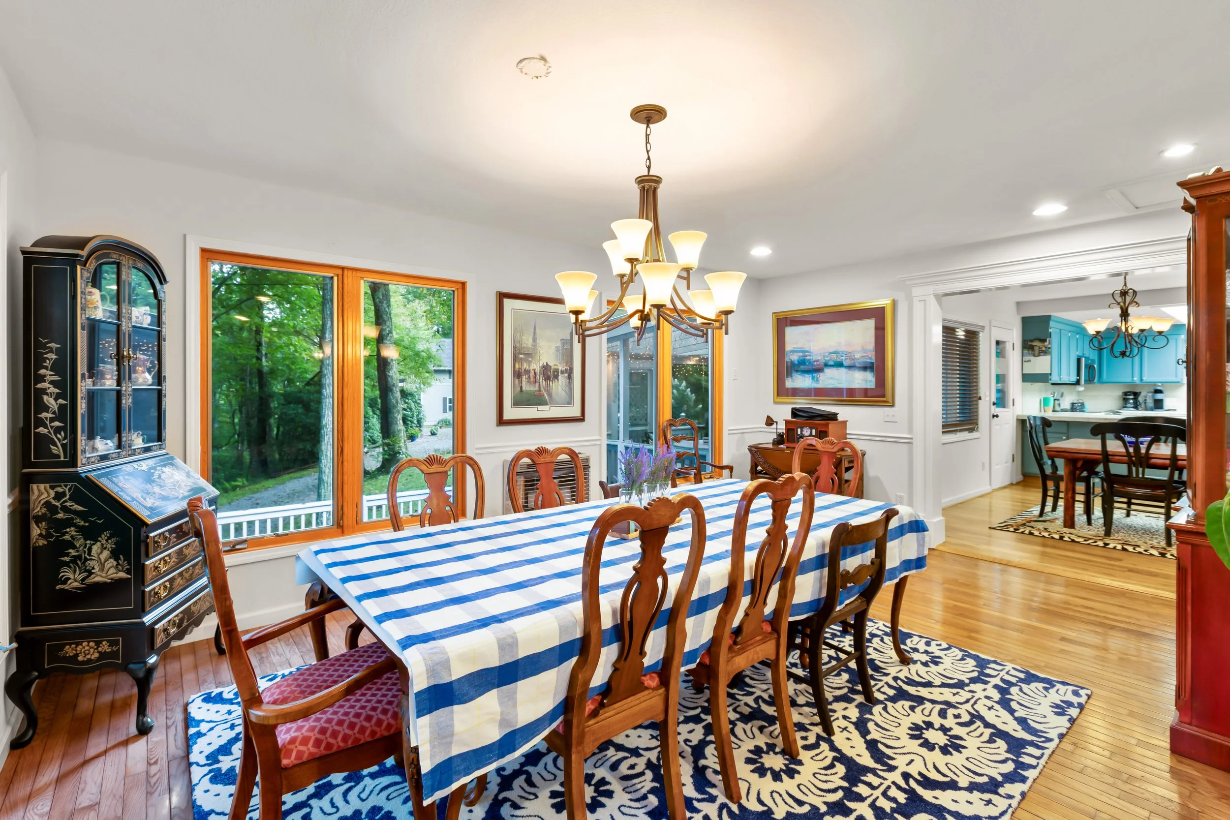 Dining room with a long table covered with a blue and white checkered tablecloth, surrounded by wooden chairs. There is a black china cabinet on the left, and large windows showing a green backyard. On the walls are framed artwork, and a chandelier h