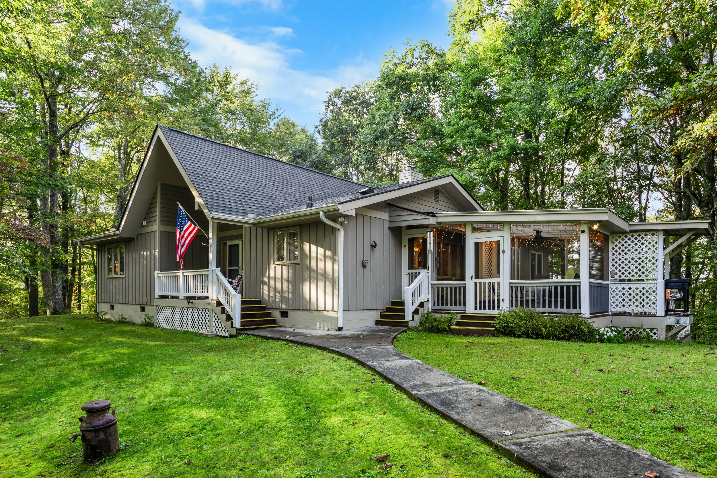 A gray house with a screened-in porch, surrounded by green lawn and trees, with an American flag on the front porch, and string lights hanging inside the screened area.