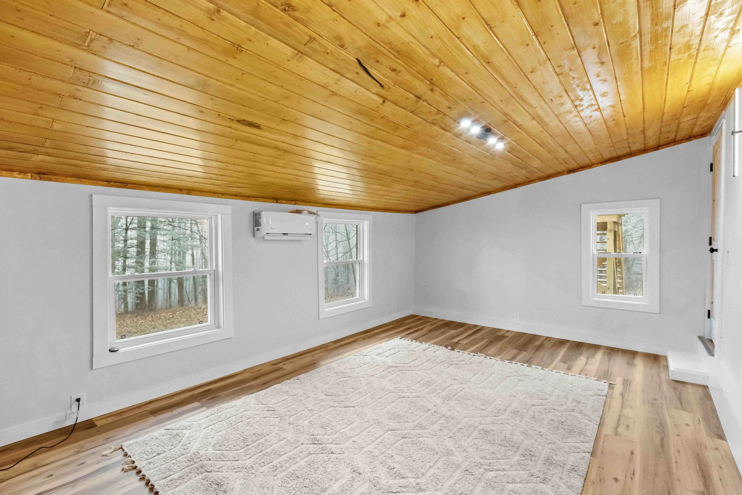 Empty room with white walls, wooden ceiling and floor, three windows, a carpet, and an air conditioning unit.