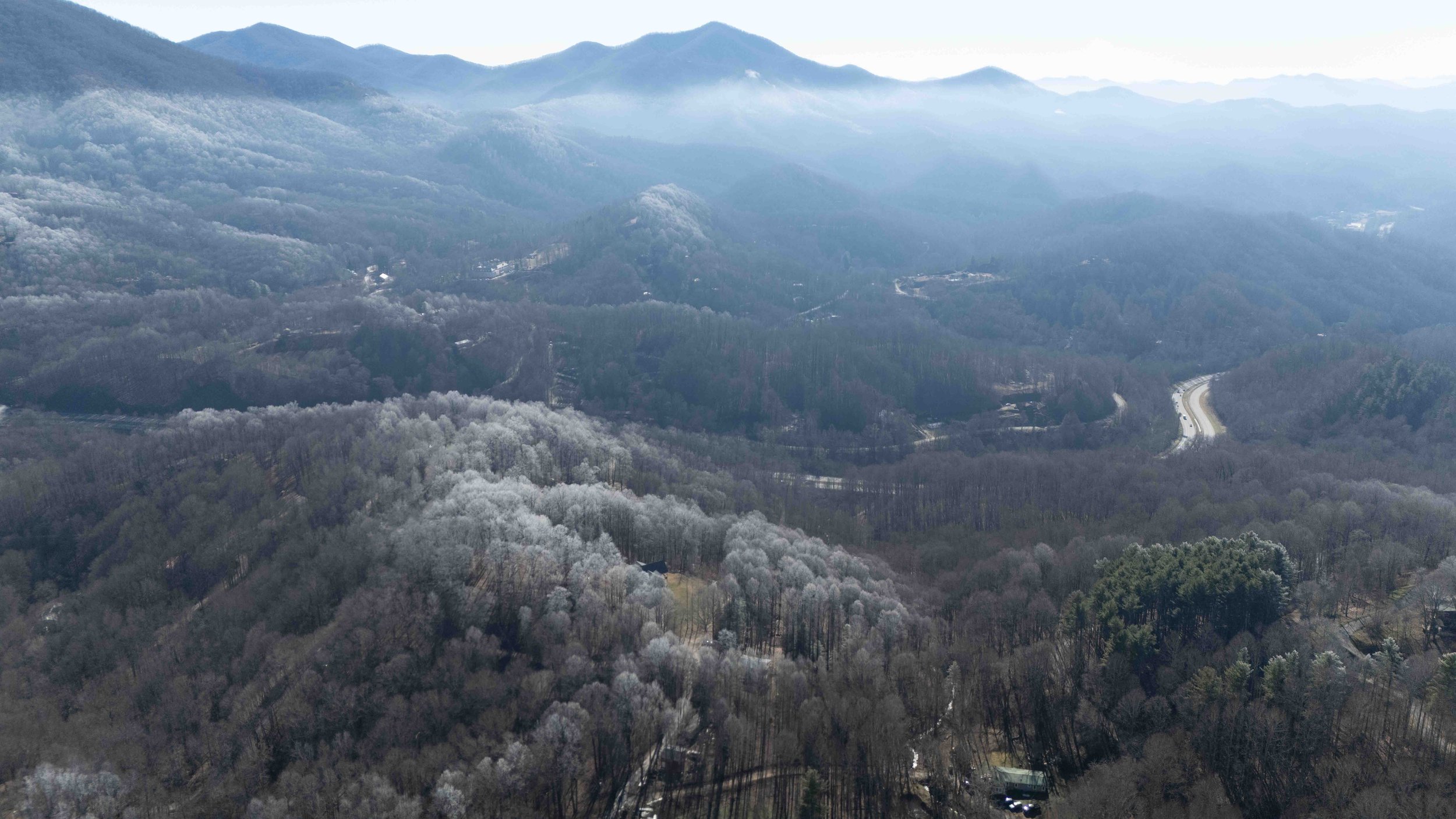 Aerial view of a mountainous landscape covered in trees with some snow, winding roads, and a hazy sky.