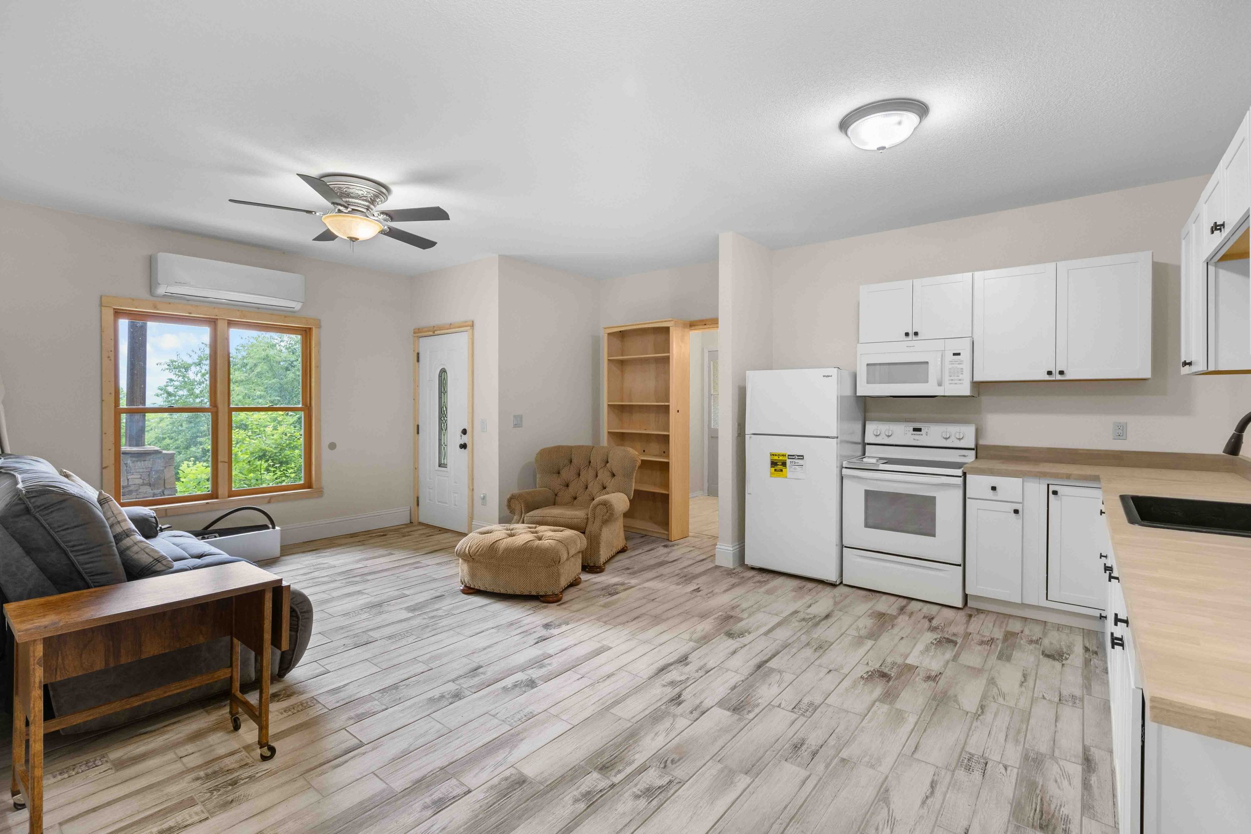 Open-concept living room and kitchen with beige walls, white cabinets, and wood flooring, featuring a window overlooking trees, a ceiling fan, and a cushioned chair.