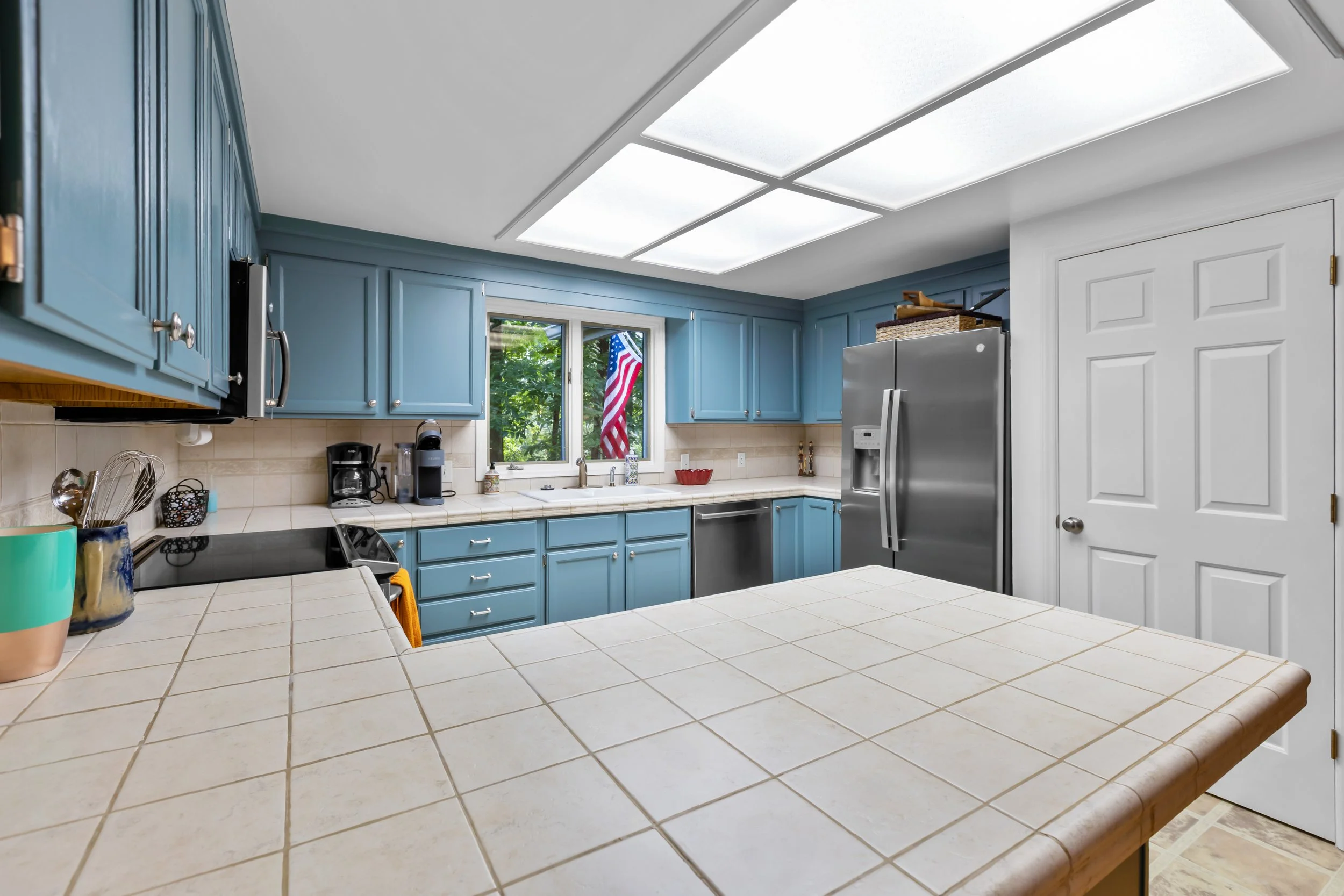 Kitchen with blue cabinets, white tiled island, stainless steel refrigerator, black stovetop, coffee maker, window with American flag, and white door.