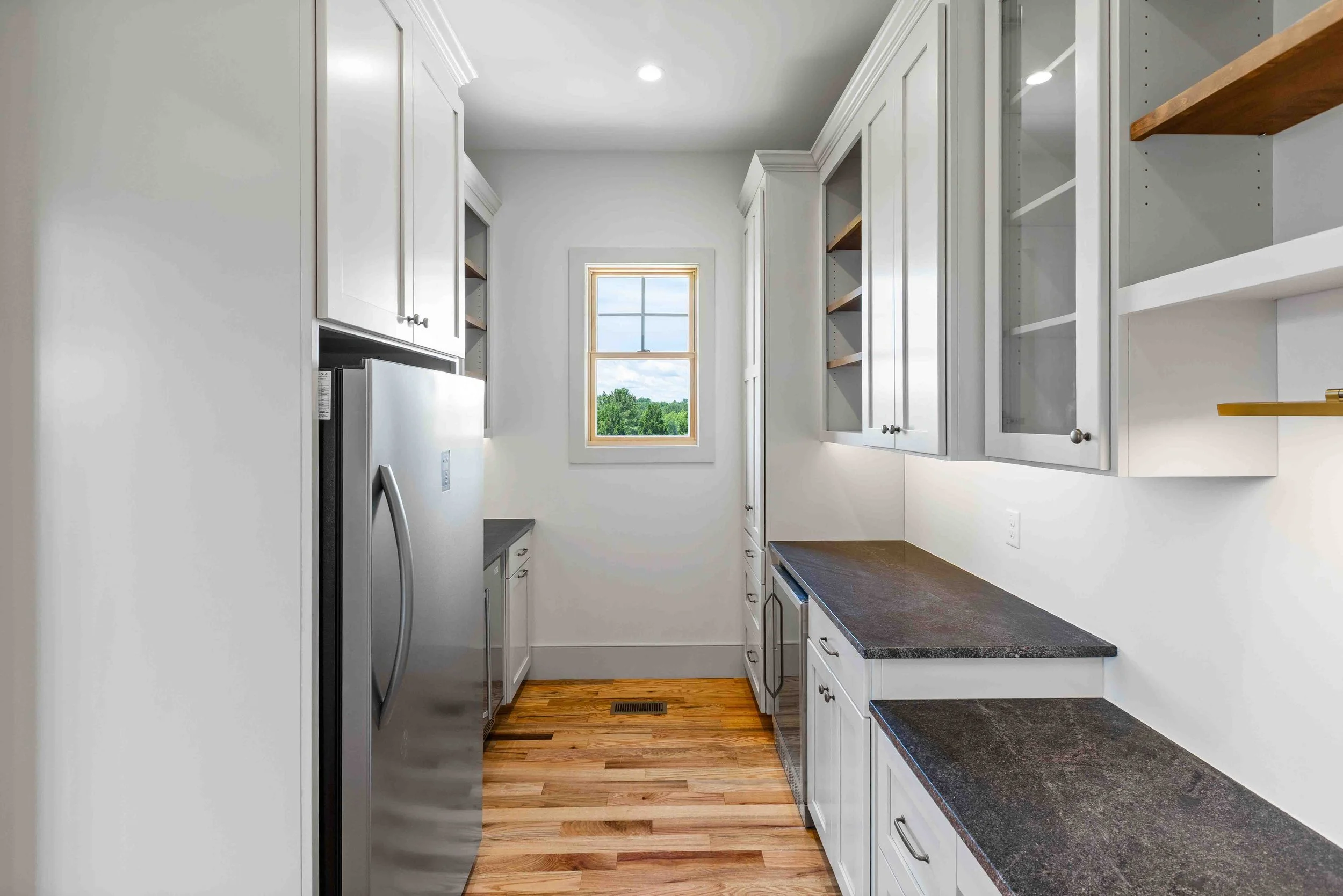 A clean, modern laundry room with white cabinets, dark countertops, a stainless steel refrigerator, and a window overlooking a green landscape.