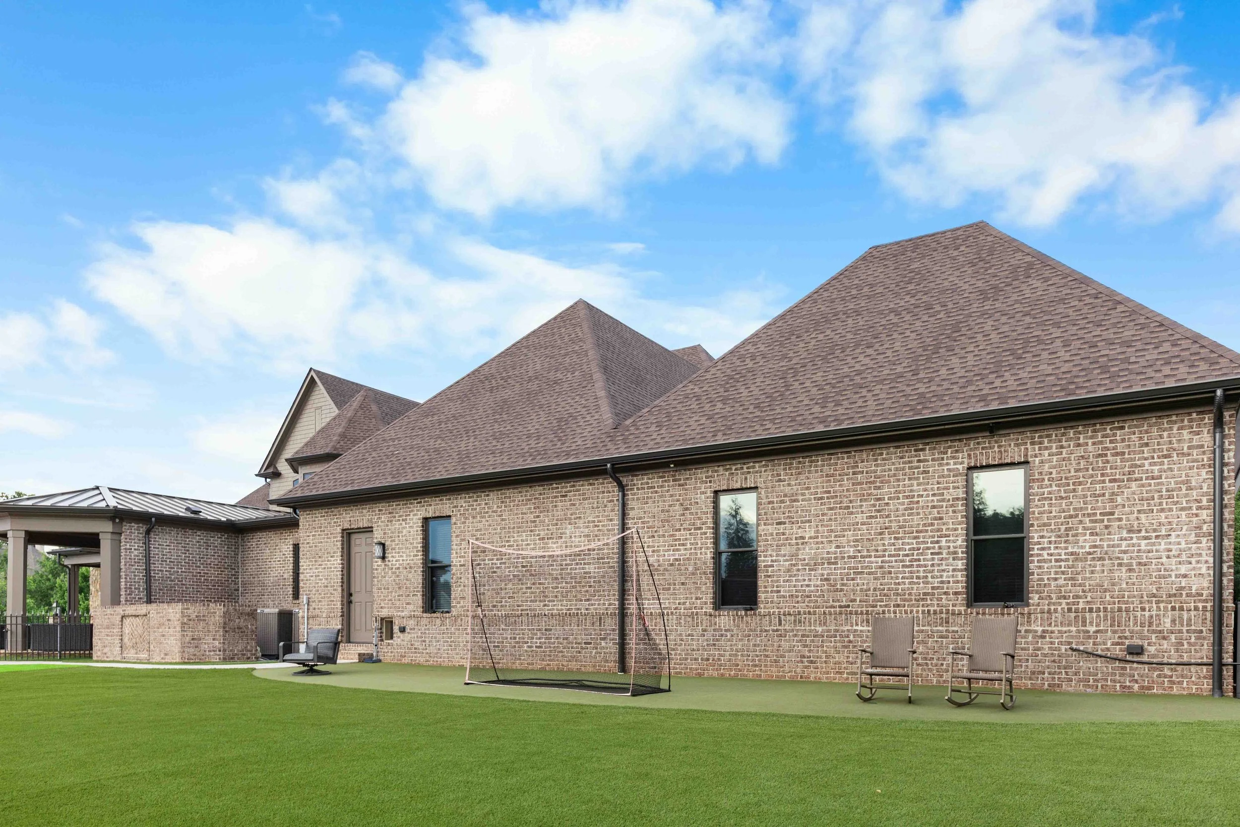 Backyard of a brick house with a green lawn, a swing set, two chairs, and a covered patio area, under a blue sky with some clouds.