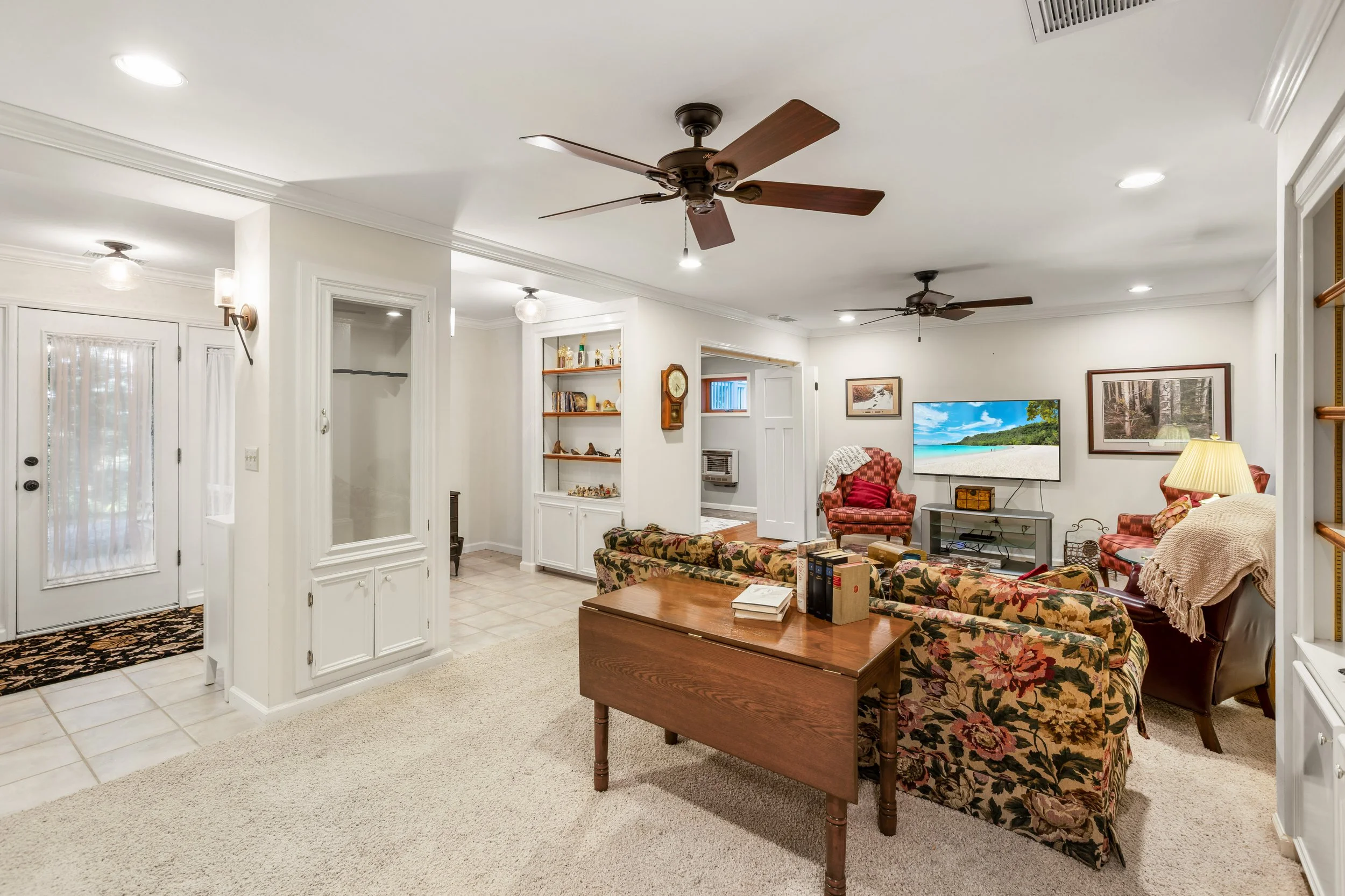 Living room with white walls, ceiling fans, wooden furniture, floral patterned sofas, a flat-screen TV, and light-colored carpet and tile flooring.