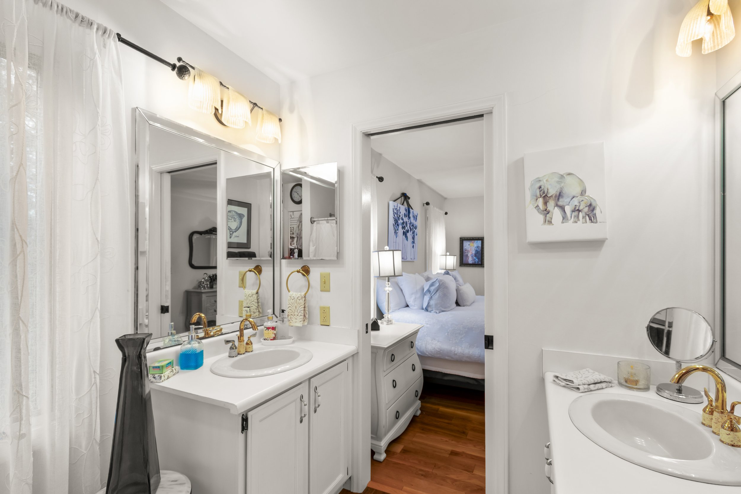 Bathroom with two sinks, mirrors, and a view into a bedroom with a bed and lamps.