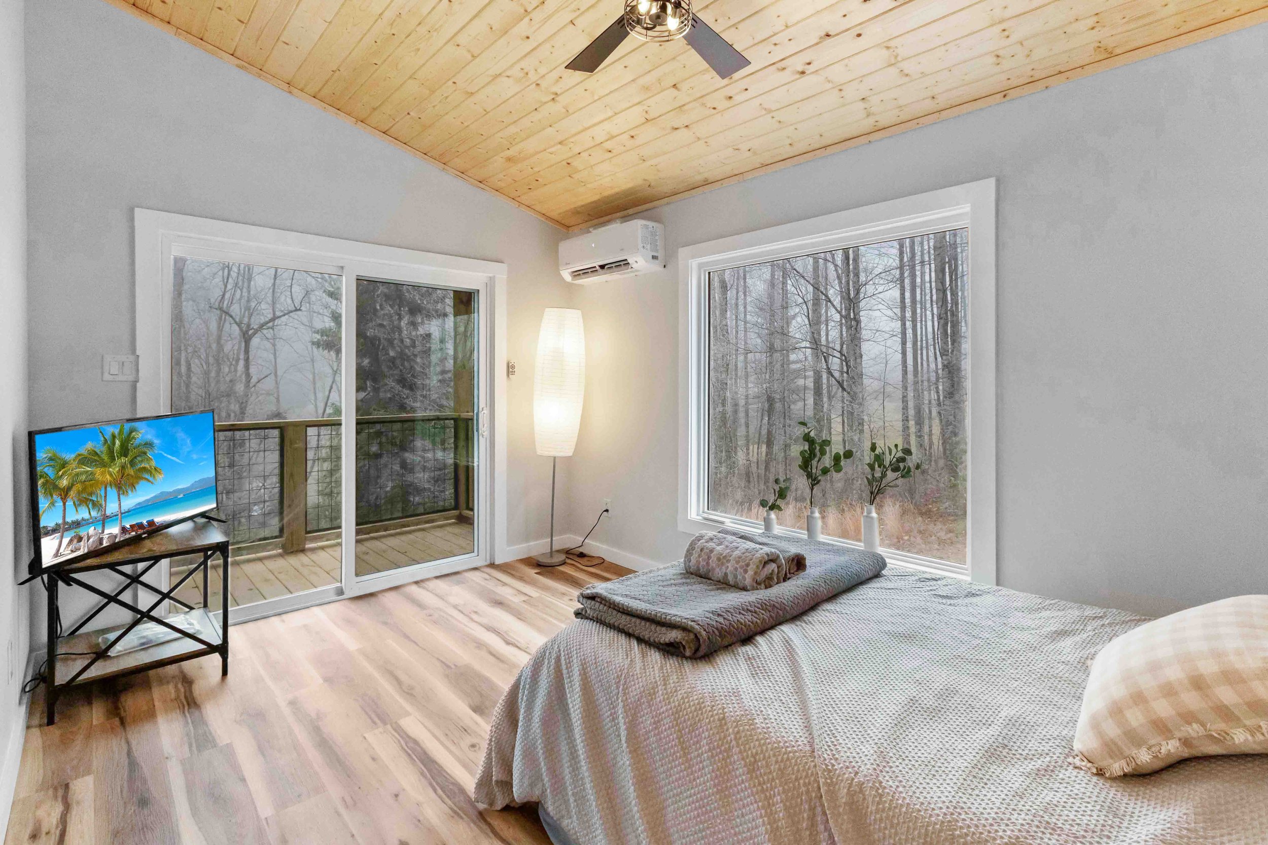 A bedroom with a wooden ceiling, gray walls, and large windows showing a forest view. The bed has beige checkered sheets, a pillow, and folded towels. There is a floor lamp, a small TV on a side table, and a sliding glass door leading to a small balc
