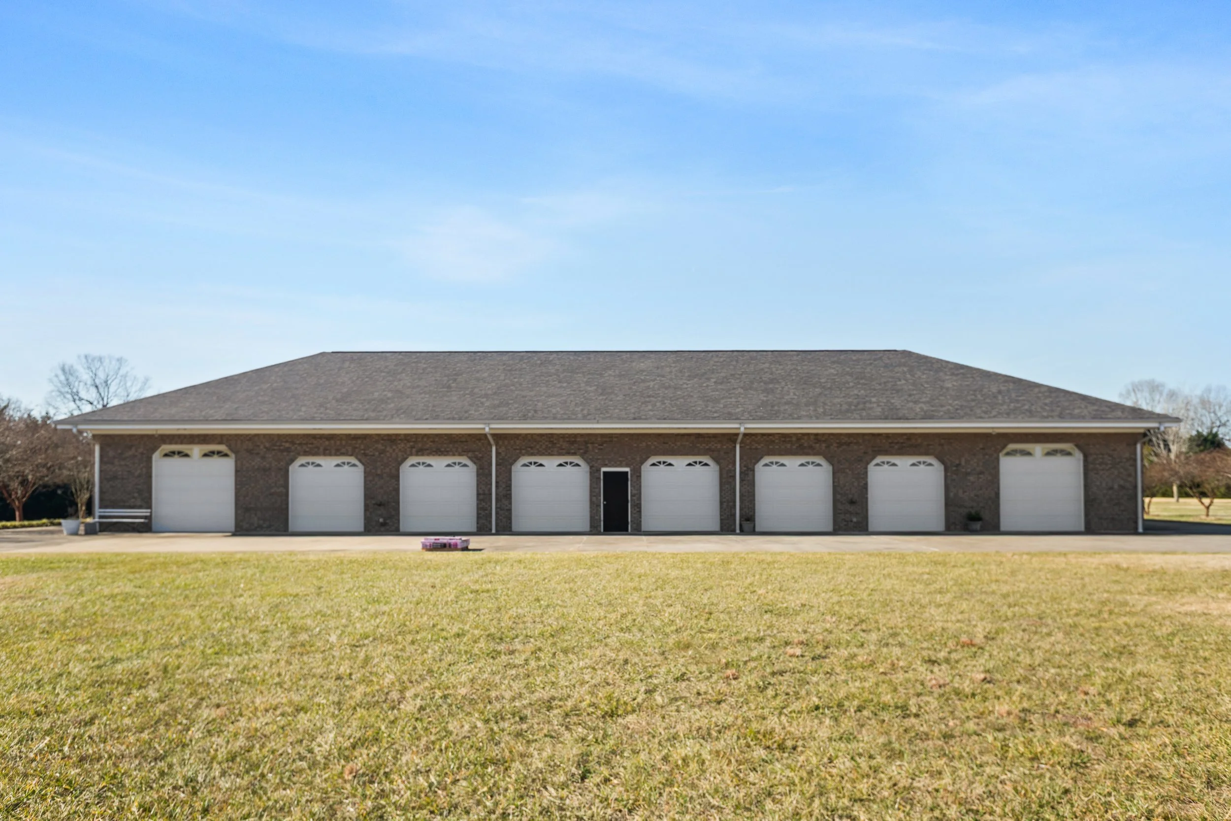 Large brick garage building with seven white garage doors and a single black side door, set against a clear blue sky and a well-maintained grassy yard.