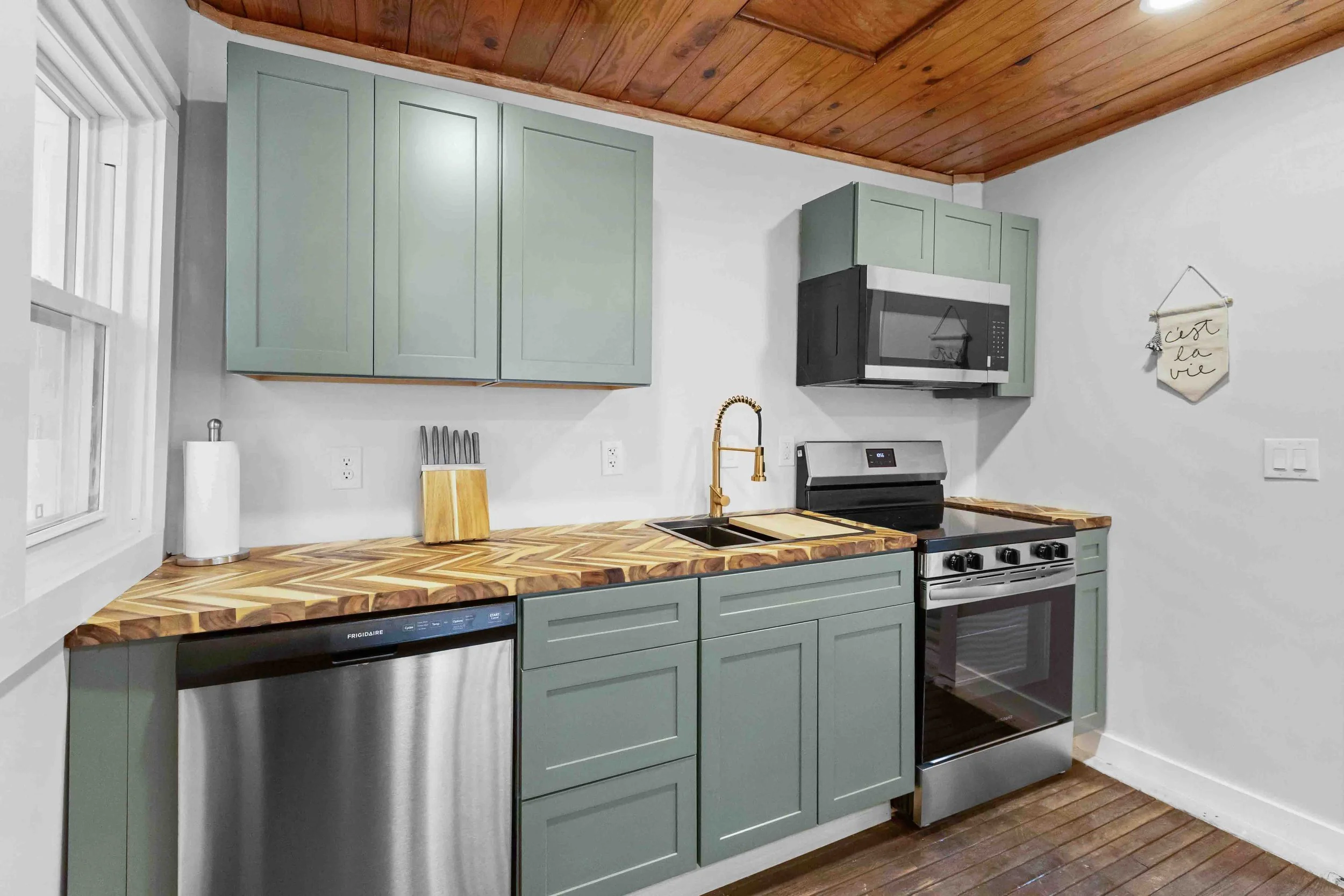Kitchen with sage green cabinets, a wood countertop, stainless steel appliances, a black microwave, oven, dishwasher, and decor including a paper towel roll, knife block, and wall hanging with French writing.