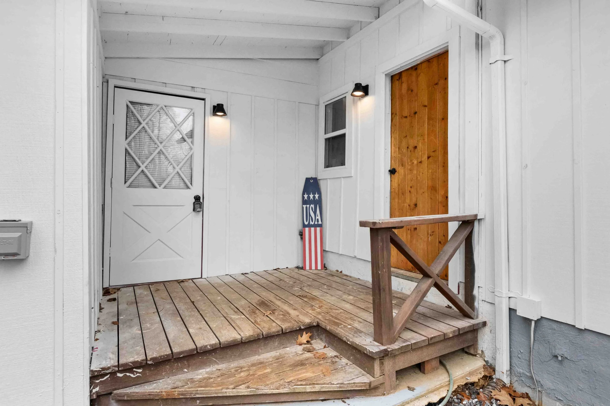 Porch area of a house with a white door with a screen and a padlock, a wooden door, and an American patriotic sign leaning against the wall. The porch has a wooden floor and a small wooden railing, with lights mounted on the wall next to the doors.