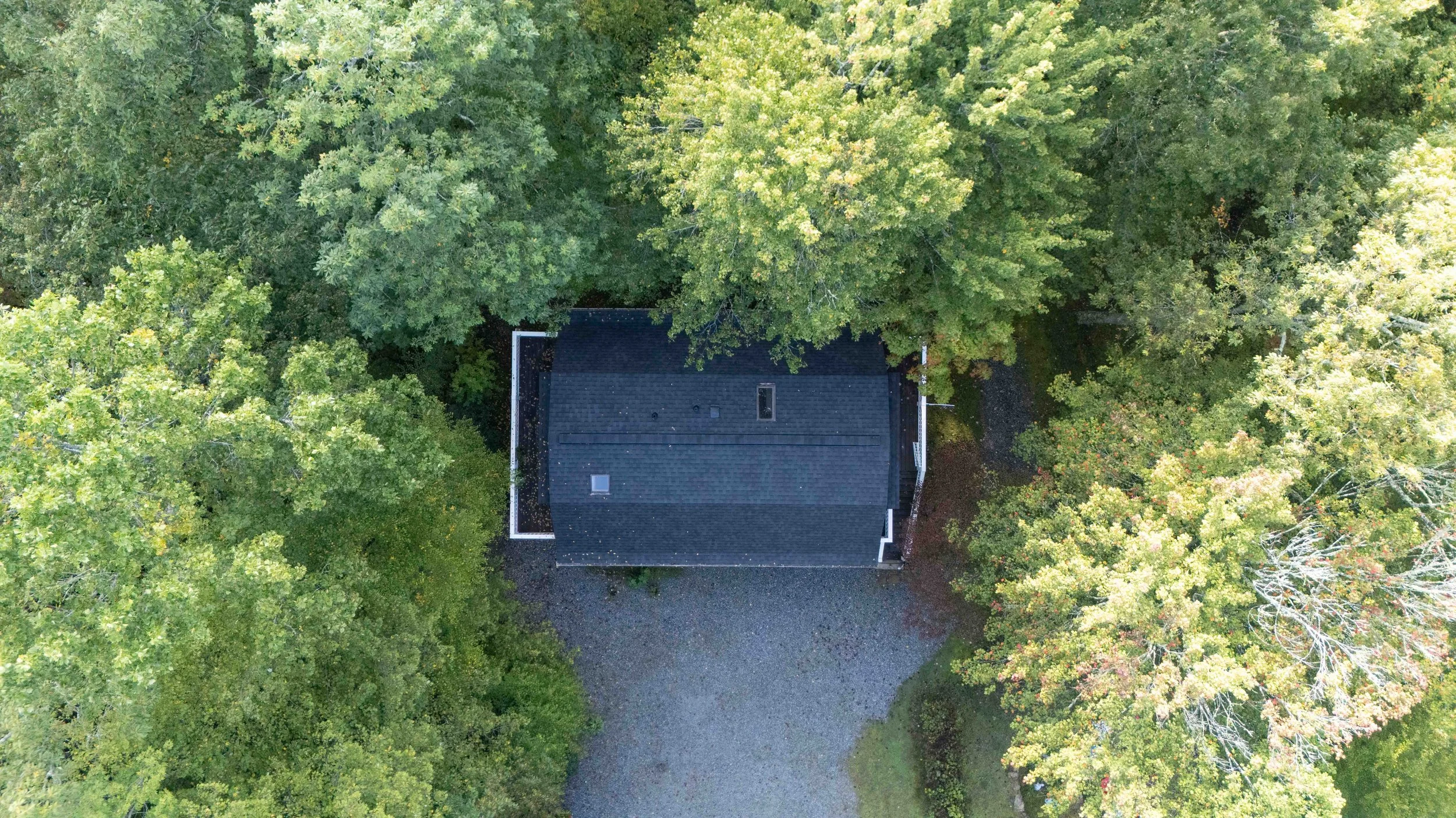 Aerial view of a house surrounded by green trees with a gravel driveway in front.